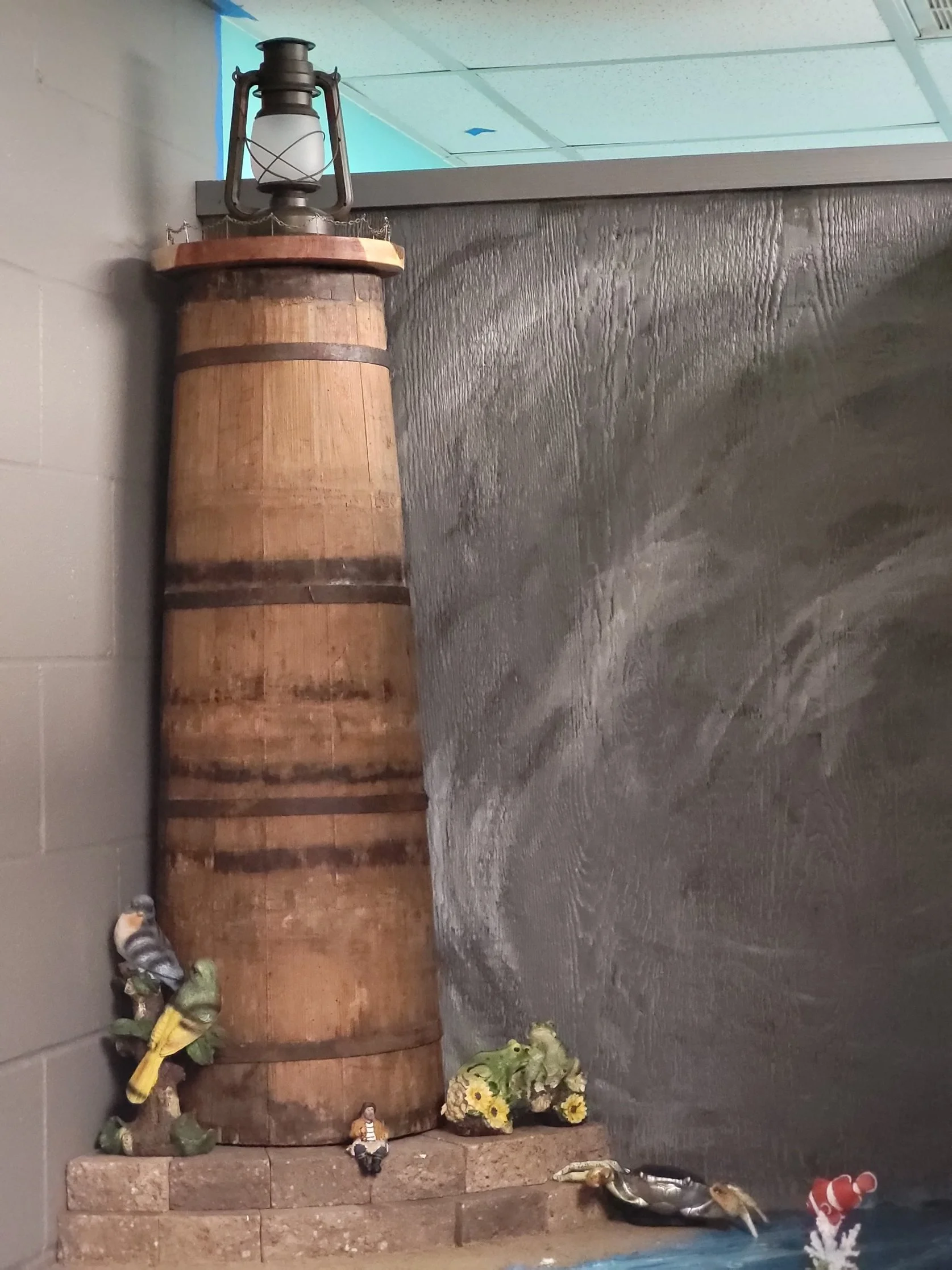 A decorative setup with a wooden barrel, vintage lantern, artificial flowers, small figurines, and a pair of shoes at the bottom on a staircase against a gray wall.