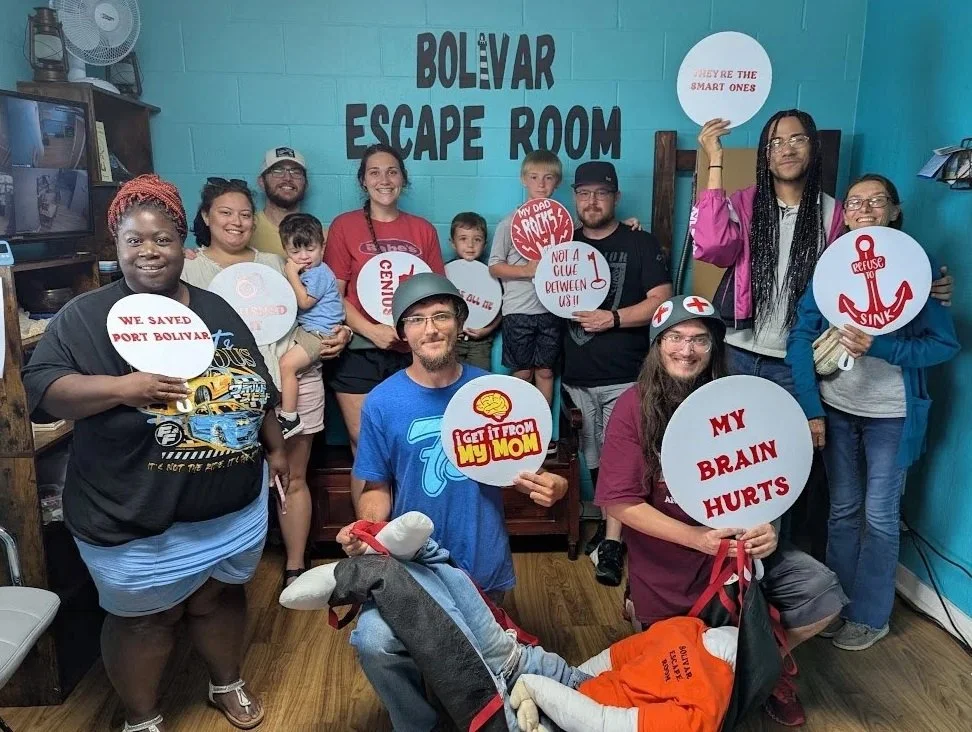 Group of people holding signs inside an escape room called Bolivar Escape Room, some signs have humorous messages like 'We saved port Bolivar,' 'My brain hurts,' and 'Refuse to sink.'