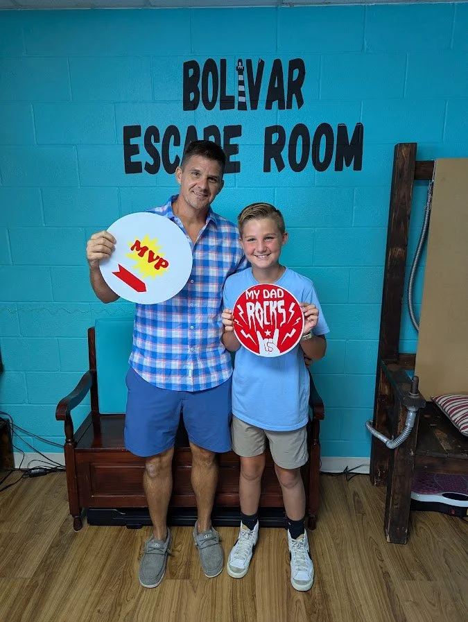 A man and boy standing in an escape room called Bolívar Escape Room, holding signs that say 'MVP' and 'My Dad Rages'.