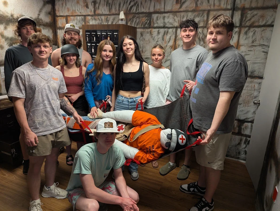 Group of nine young people standing around a person lying on a stretcher in a room with textured walls, with some holding the stretcher's straps. The person on the stretcher is dressed as a rescue worker with orange gear, a hat, and sunglasses, and h