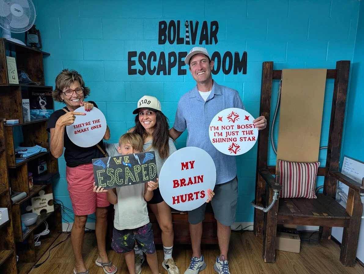A group of four people, including one child, standing inside an escape room called 'Bolivar Escape Room.' They are smiling and holding signs with humorous messages about their experience. The background features a bright blue wall, a wooden bookshelf