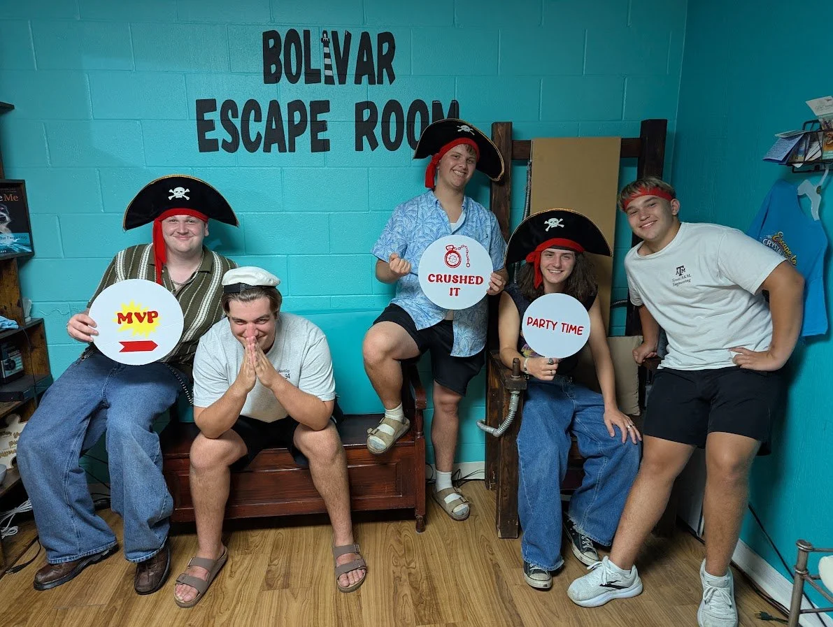 Five young people dressed as pirates, some with pirate hats, in a room with teal walls and a sign that says "Bolivar Escape Room". They hold signs and pose for a photo, smiling and making playful gestures.