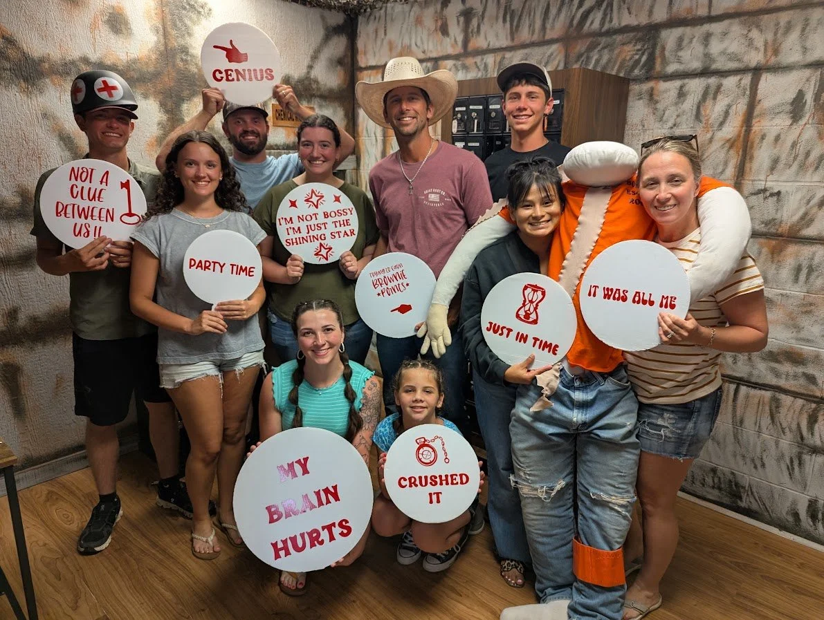 Group of people posing with humorous signs and costumes in a room with wooden floors and a rustic wall. Some wear costume accessories like a nurse hat, cowboy hat, and a crocodile costume. The signs include phrases like "Not a clue between us," "Part