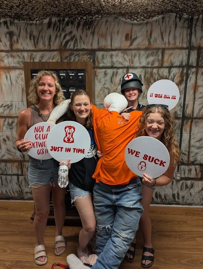 Four women and one person dressed as a soldier play with signs with funny and rebellious phrases in an indoor setting with graffiti-style walls.