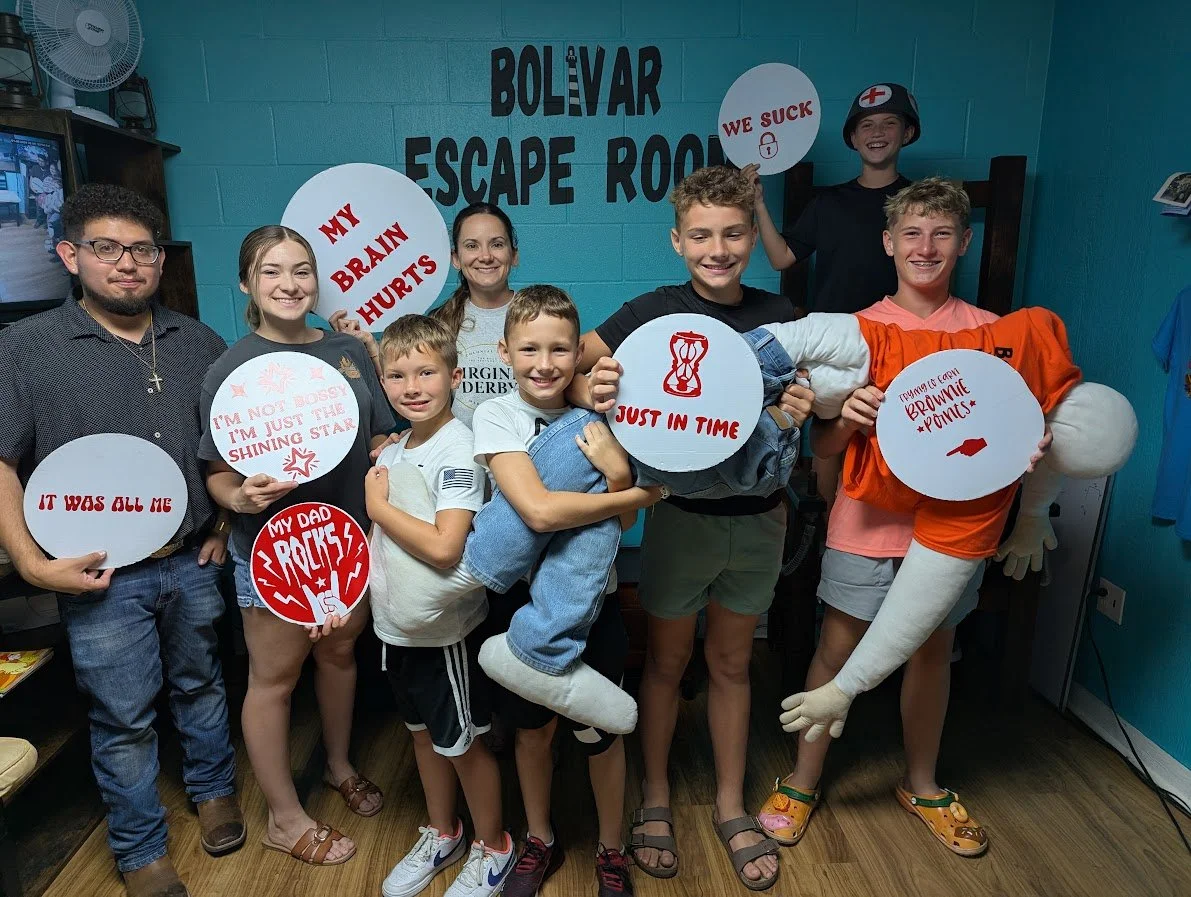 Group of children and a woman in an escape room, holding signs with humorous messages and a mannequin dressed as a nurse. The background displays a sign reading 'Bolivar Escape Room'.