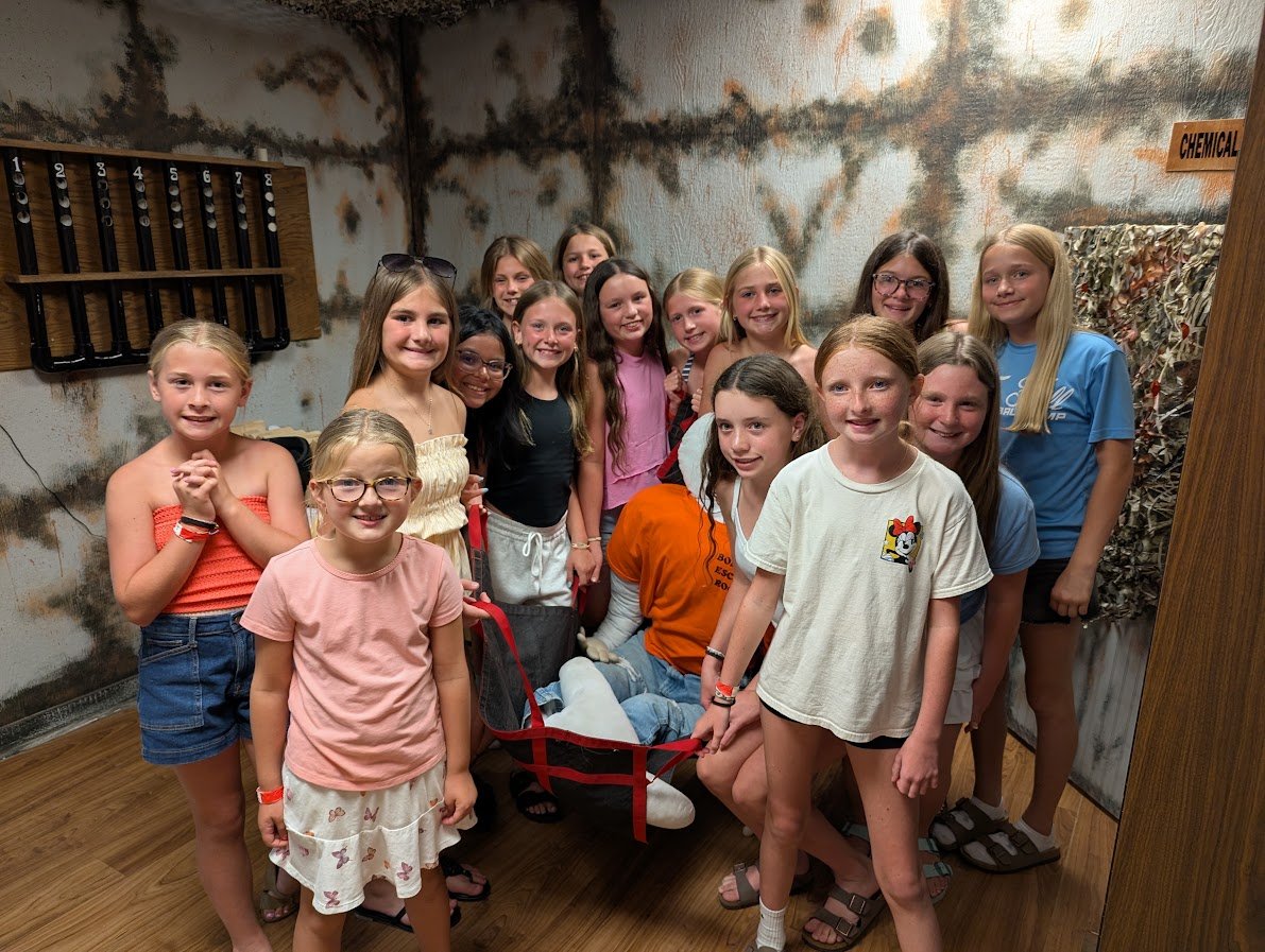 Group of children dressed in casual summer clothes, surrounding a person lying in a faux rescue stretcher, at an indoor facility with a wall sign reading 'Chemical' and textured walls.