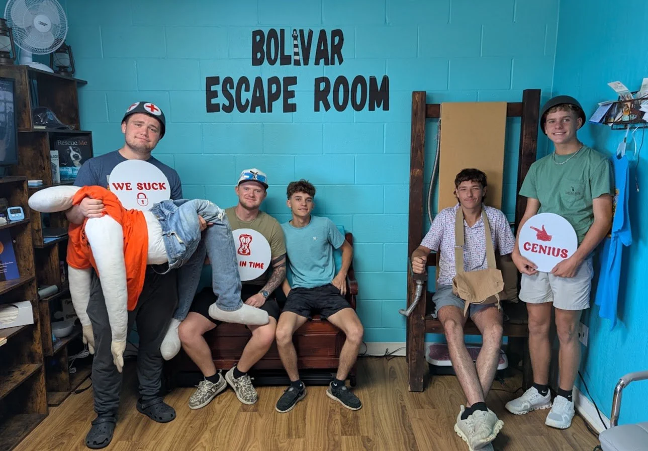 Five young men inside an escape room, with a blue wall behind them that reads "Bolvar Escape Room." One man on the left is dressed as a nurse, holding a person in a costume. The others are sitting or standing, holding signs with phrases like "We suck