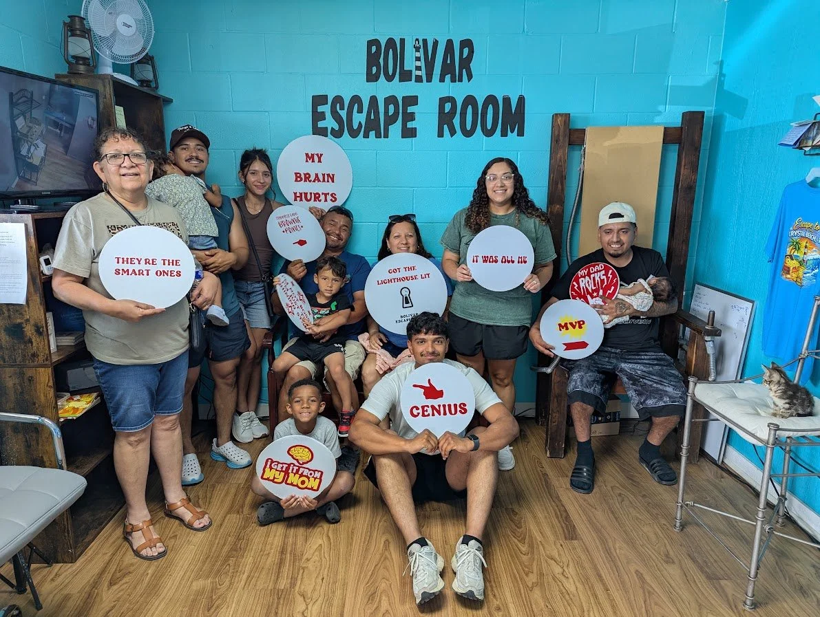 Group of people celebrating after escaping from an escape room titled 'Bolivar Escape Room.' They are holding signs with playful messages like 'Genius,' 'It was all me,' 'My dad rocks,' 'MVP,' 'They’re the smart ones,' and 'My brain hurts.' Some are 