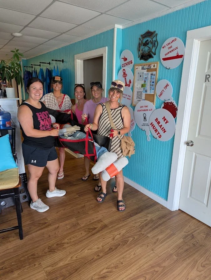 Five women standing in a room with light blue walls, some holding a golf bag with clubs, and surrounded by themed signs on the wall. The women are smiling and dressed casually for a summer outing or party.