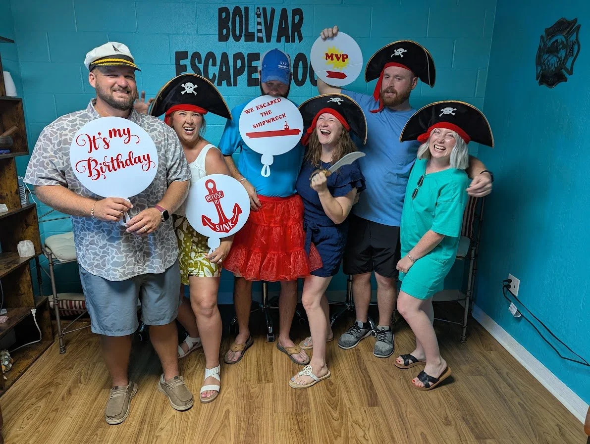 Group of six people dressed as pirates with hats, holding signs for a birthday celebration at an escape room, with blue walls and wooden flooring.