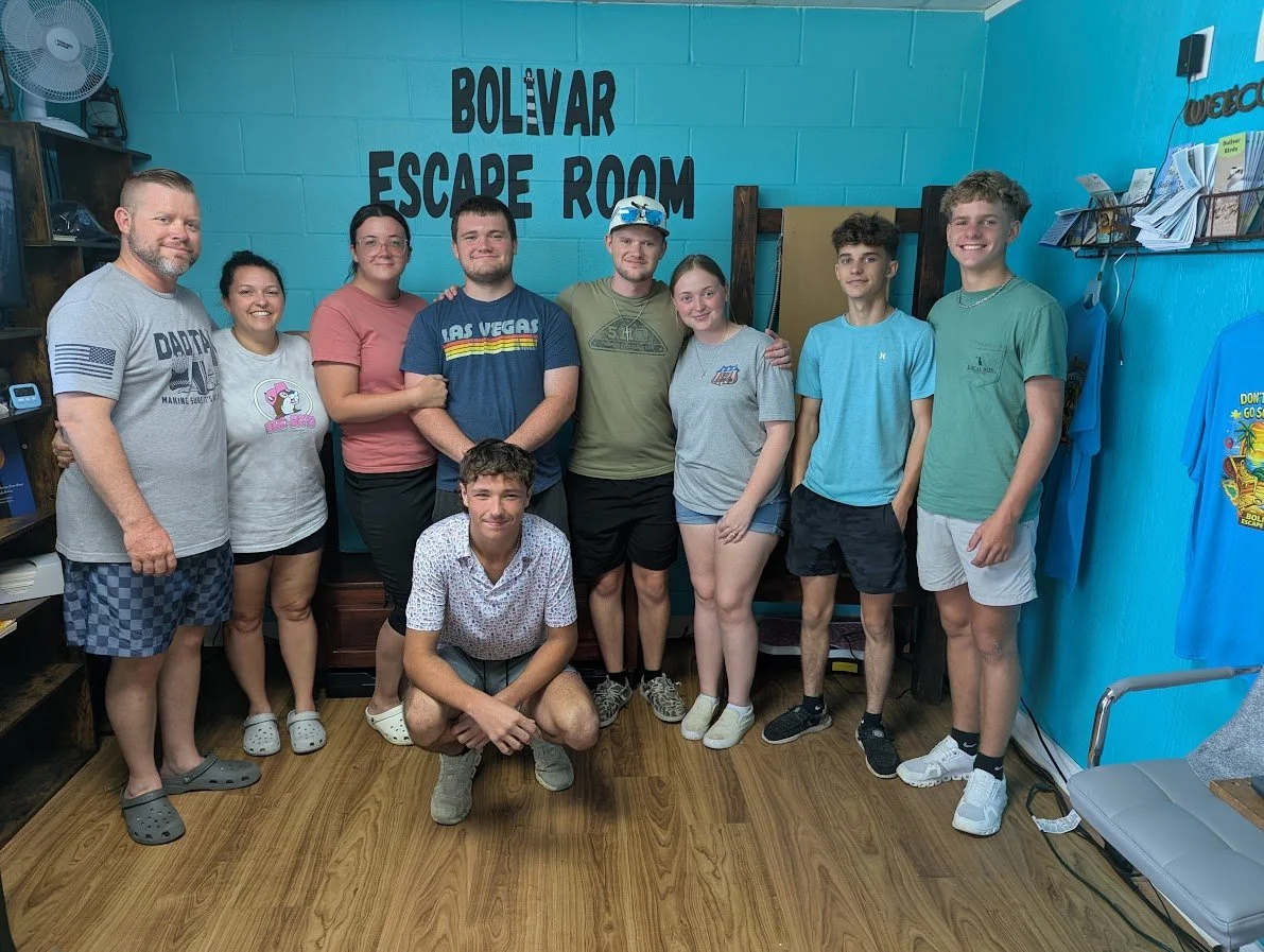 Group photo of ten young people and two adults standing inside a room with blue walls, branded as 'Bolvar Escape Room', smiling at the camera, some with arms around each other, with bookshelves and escape room decorations in the background.