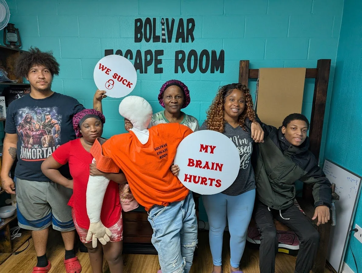 Group of six people, including children and adults, standing in a room labeled 'Bolvar Escape Room'. One person dressed as a guard is holding signs that say 'We Suck' and 'My Brain Hurts'. The group appears to be participating in or celebrating an escape room activity.