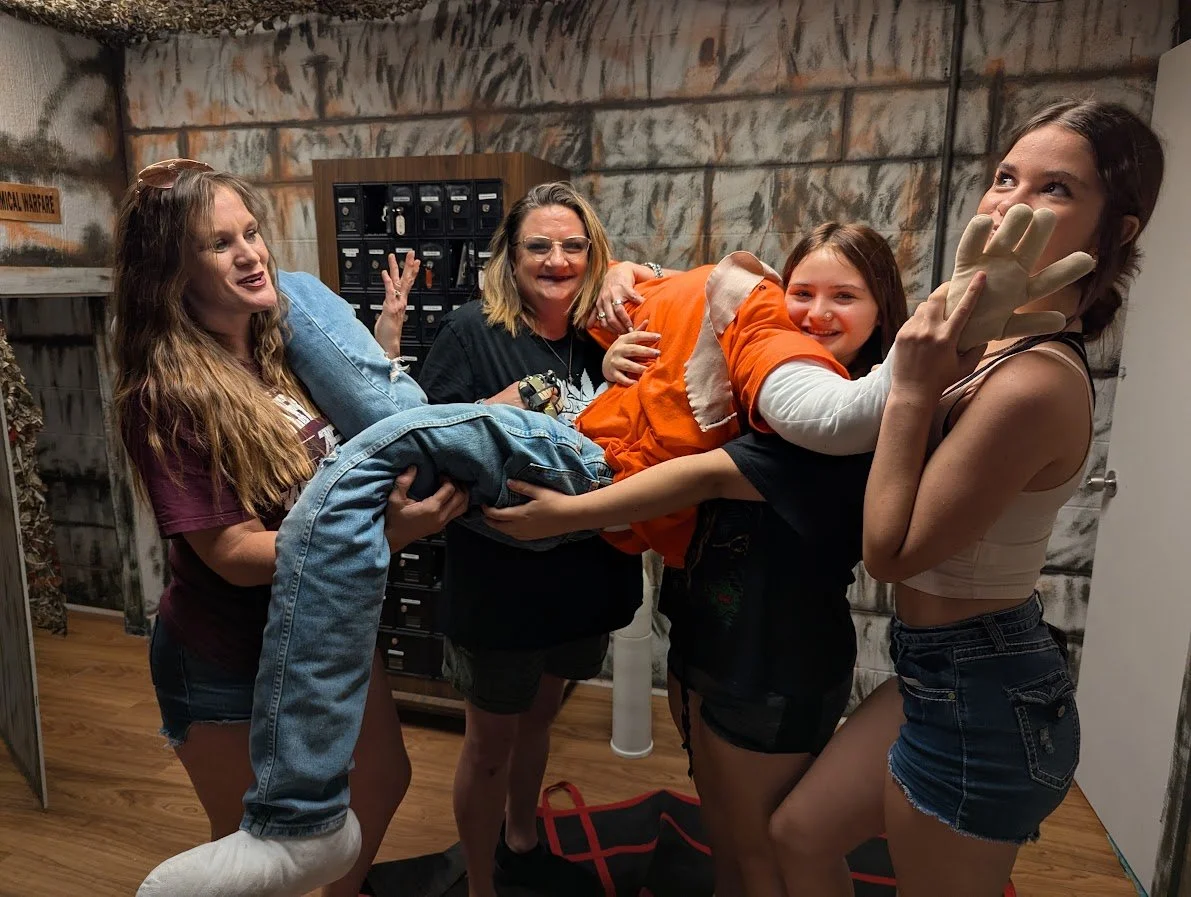 Group of five women and girls smiling and holding a person in a costume, with a dark, textured wall, a wooden cabinet, and a sign that reads 'Chemical Warfare' in the background.
