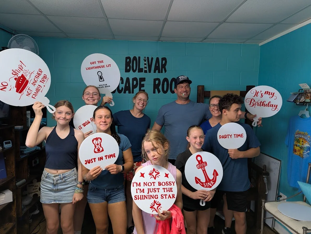 Group of children and adults celebrating inside Bolivar Escape Room, holding themed signs with phrases like 'No Shrugging! Not a family trip,' 'Got the lighthouse lit,' 'It's my birthday,' and 'Party time.'