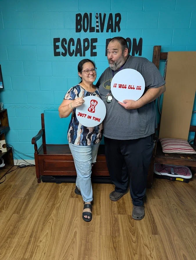 Two people standing in front of a blue wall with text reading 'Bolivar Escape Room.' They are holding round signs with red lettering; the woman’s sign says 'Just In Time' with a graphic of a dress, and the man's sign says 'It Was All Me.' Both are sm