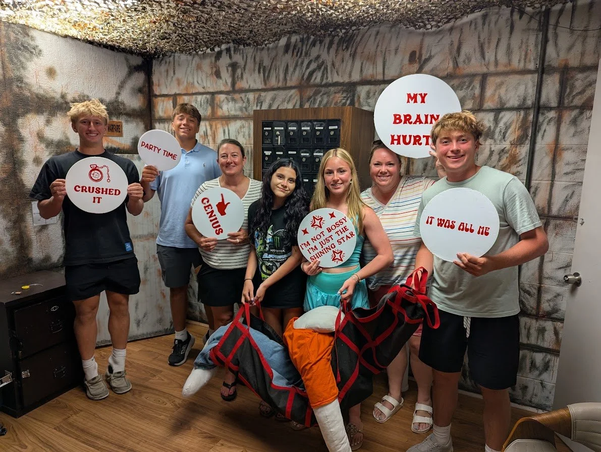 Group of seven people holding signs with various phrases, standing in front of a graffiti-style wall inside a room, with a mountain of gear in front of them.