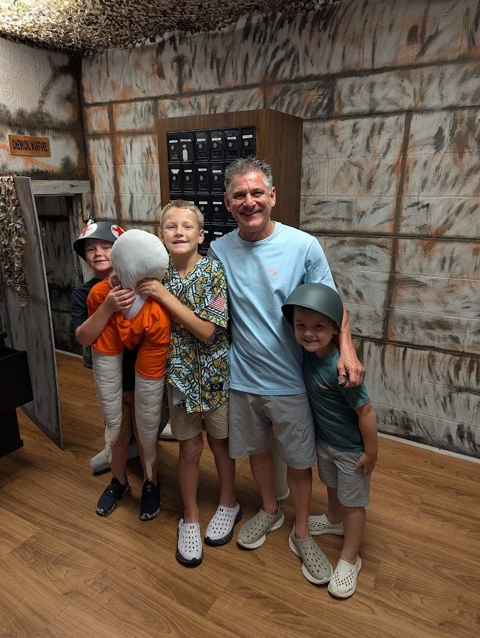 Four children and an adult man dressed as a marine, smiling and standing together in a room, with the children wearing makeshift costumes resembling a sailor, a submarine, and a military helmet.