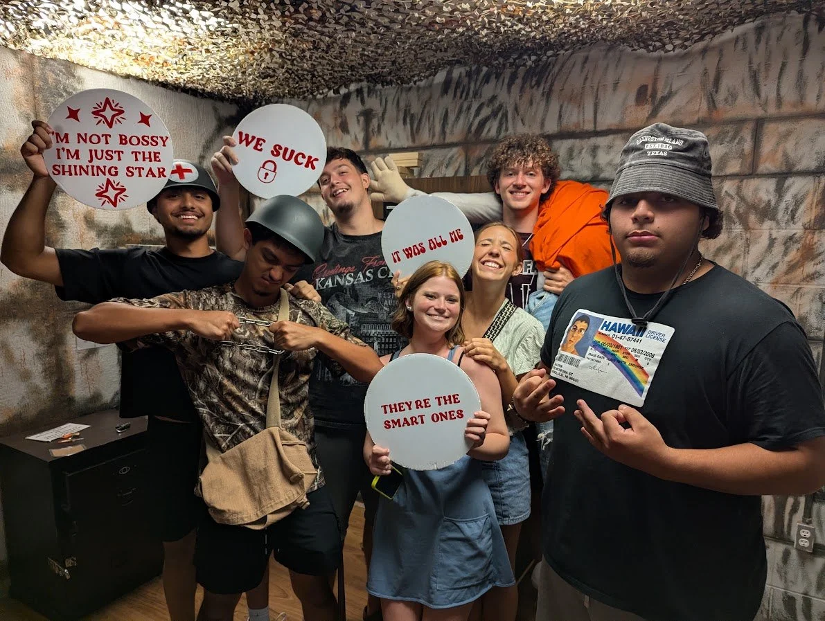Group of seven young adults smiling and posing together in an indoor space with graffiti-style walls. Some hold signs with playful messages, and one wears a military helmet, while another has a 'Hawaii' ID badge with a rainbow flag.