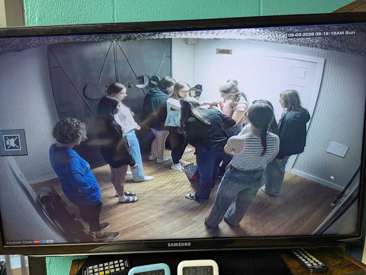 A group of people standing inside a room, possibly in a detention or waiting area, labeled 'BARRACKS.' Some individuals are engaged in a discussion while others are waiting in line. The room has wood flooring, a white door, and a gray wall with geome
