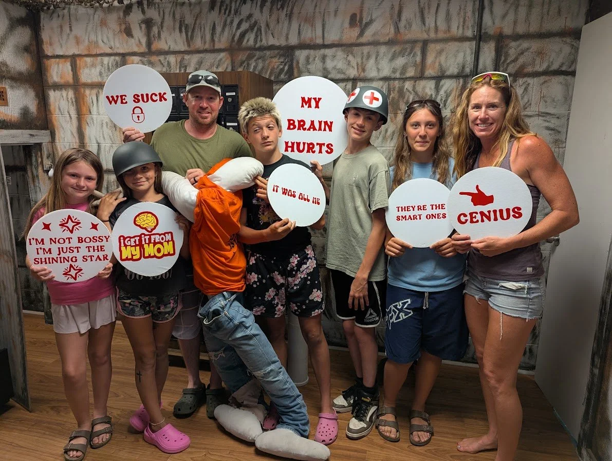 Group of eight people, including kids and two adults, holding signs with humorous messages. The signs read: "WE SUCK," "MY BRAIN HURTS," "I'M NOT BOSSY I’M JUST THE SHINING STAR," "GET IT FROM MY MOM," "IT WAS ALL ME," "THEY'RE THE SMART ONES," and "