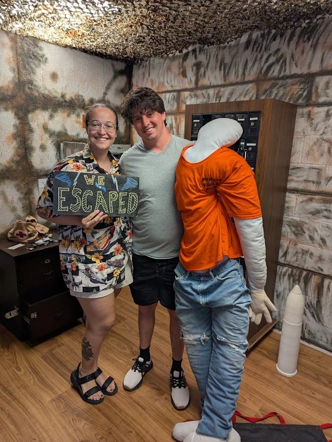 Two smiling young adults, a man and a woman, standing with a humanoid robot in a room with wooden floors and graffiti-covered walls. The woman is holding a sign that says "We Escaped" and is wearing a floral shirt and shorts. The man is wearing a gra
