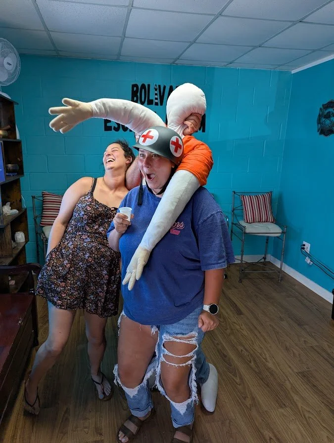 Two women and a person in a costume. The woman on the left is laughing and wearing a floral dress. The woman in the center is holding a small object and wearing a gray shirt with distressed jeans. The person in the costume is wearing a nurse hat with a red cross, gloves, and has a large, head mask with an exaggerated arm reaching around the woman's neck. They are in a room with blue walls and wooden floors, with chairs and a shelf in the background.
