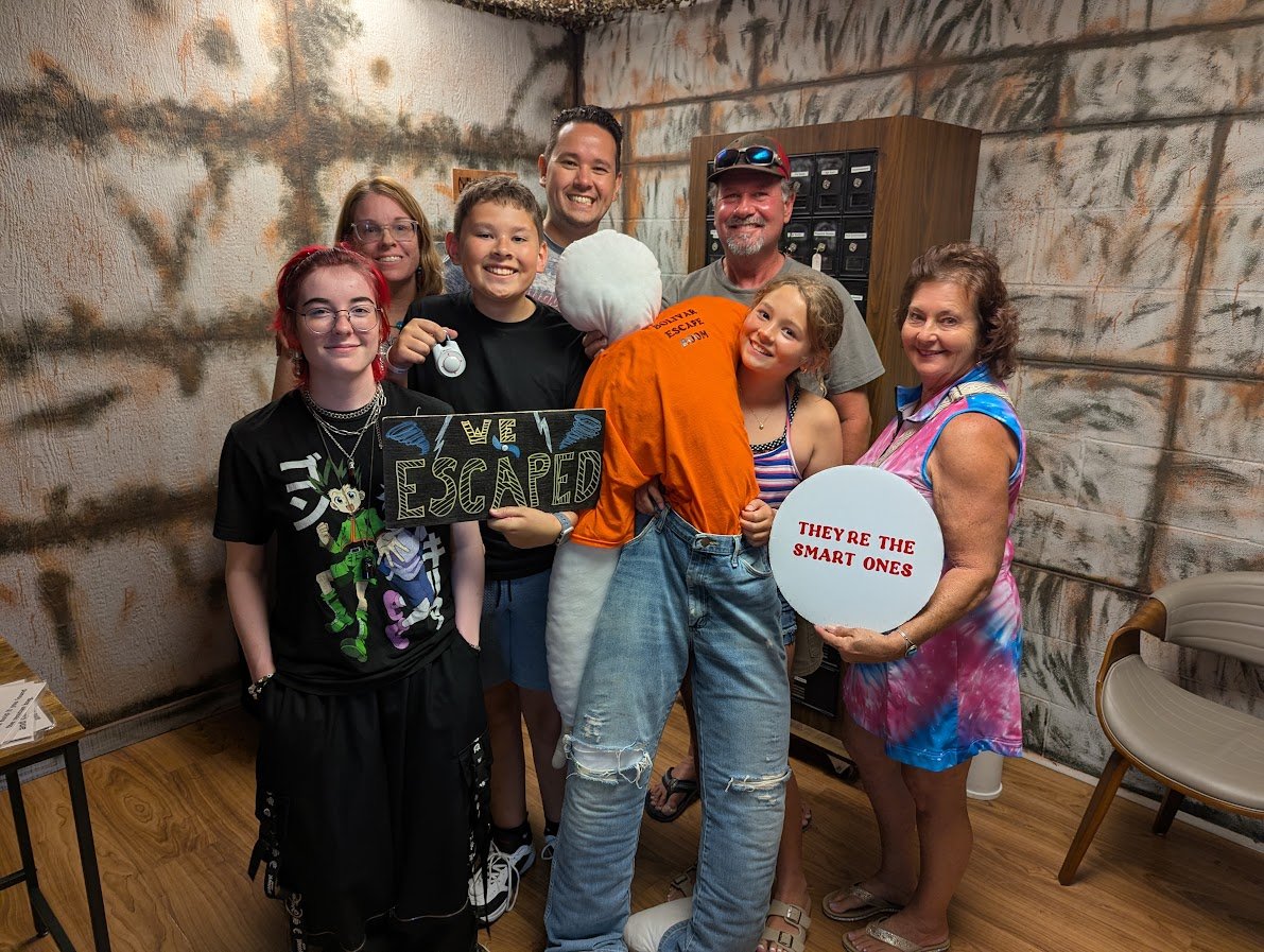 A group of people celebrating an escape room victory, holding signs that say 'WE ESCAPED' and 'THEY'RE THE SMART ONES'. One person is dressed in an orange inmate costume. Everyone is smiling and posing in a room with concrete wall decor and mailboxes
