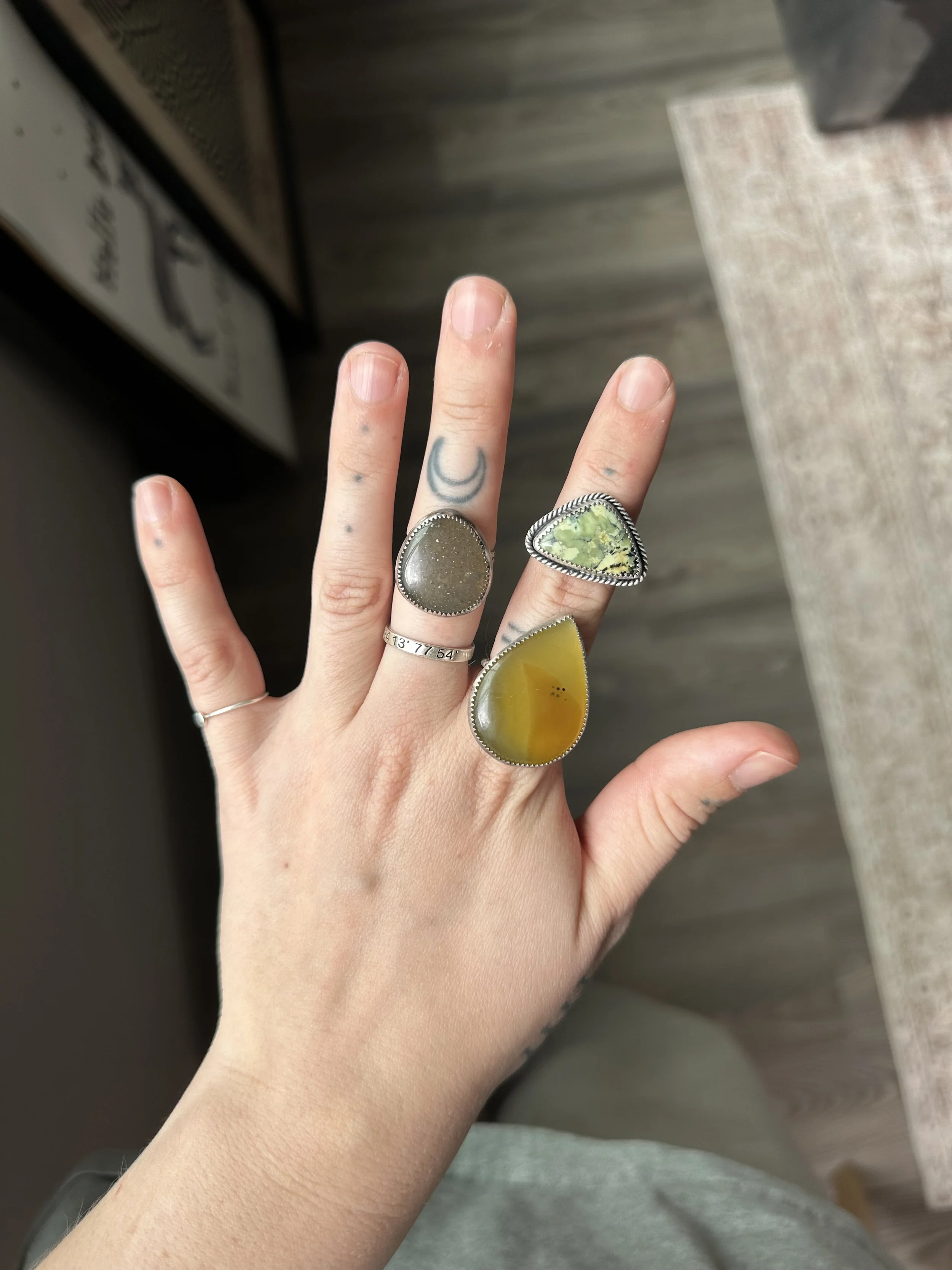 A person's hand with multiple rings, showing fingers with some tattoos, and three large rings with different stones, held near a wooden surface.