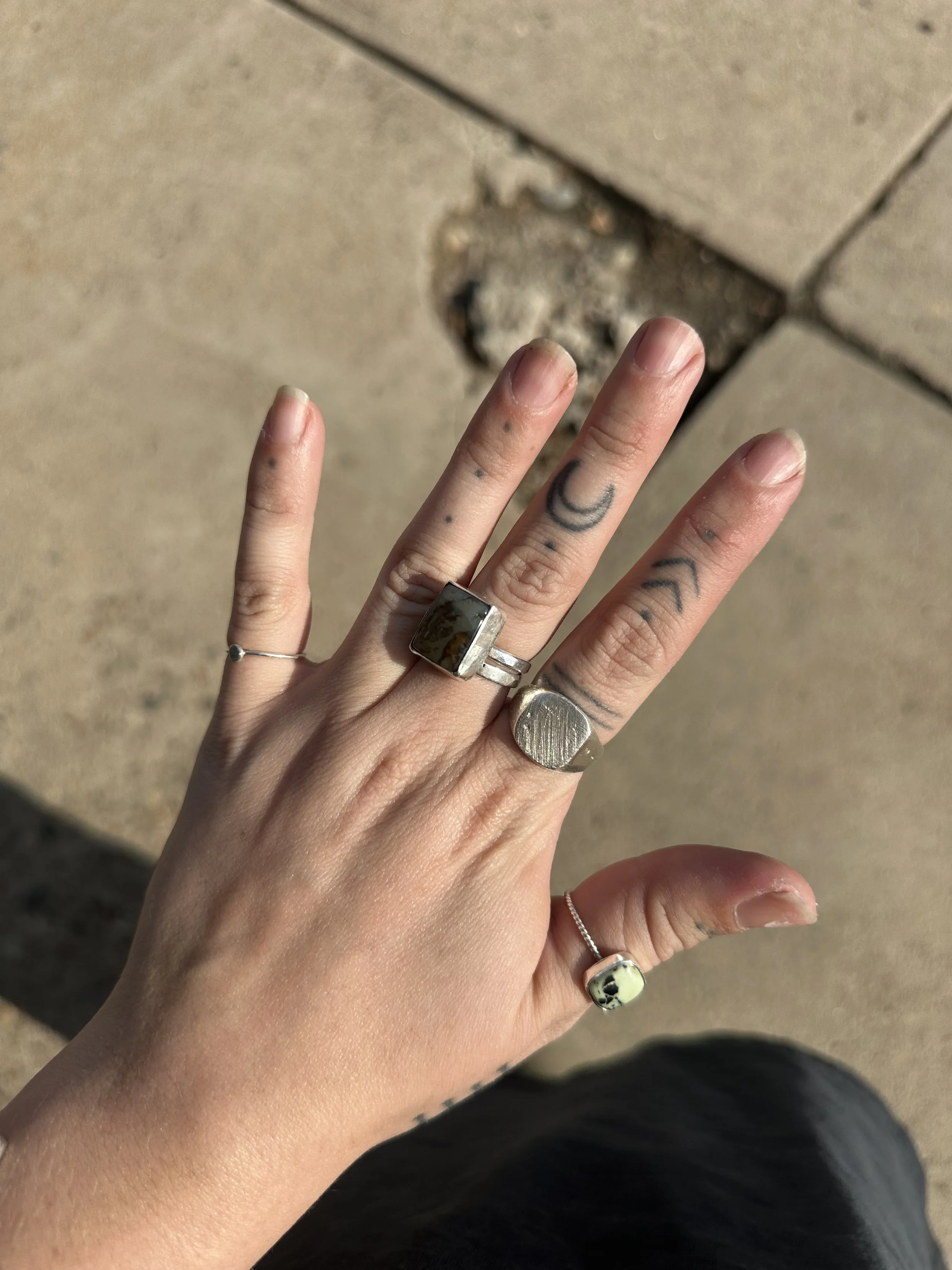 A person's hand displaying multiple rings and tattoos, with a background of beige tiled flooring.