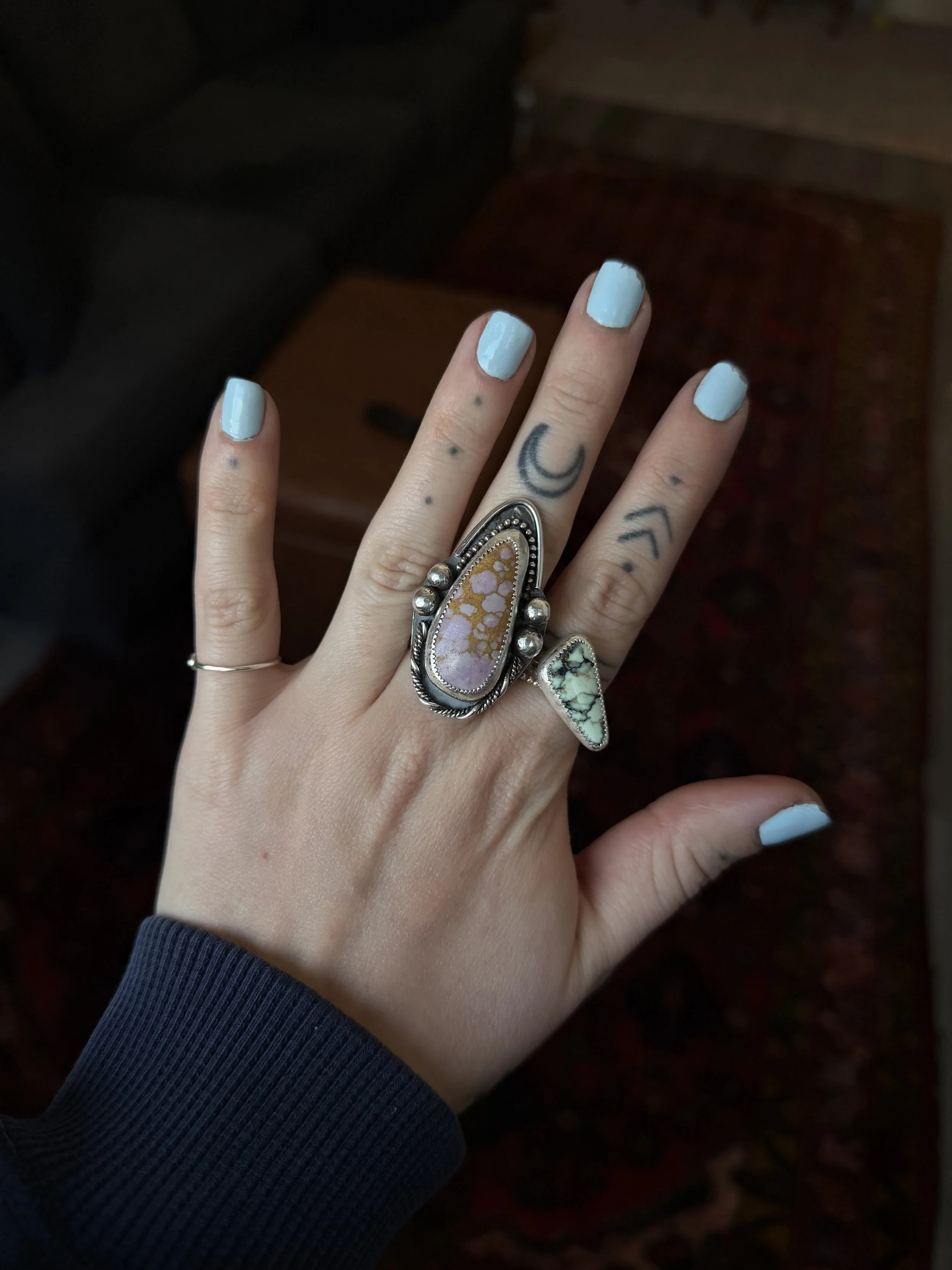 Hand with light blue painted nails wearing several rings, including a large turquoise and silver statement ring, against a dark background.