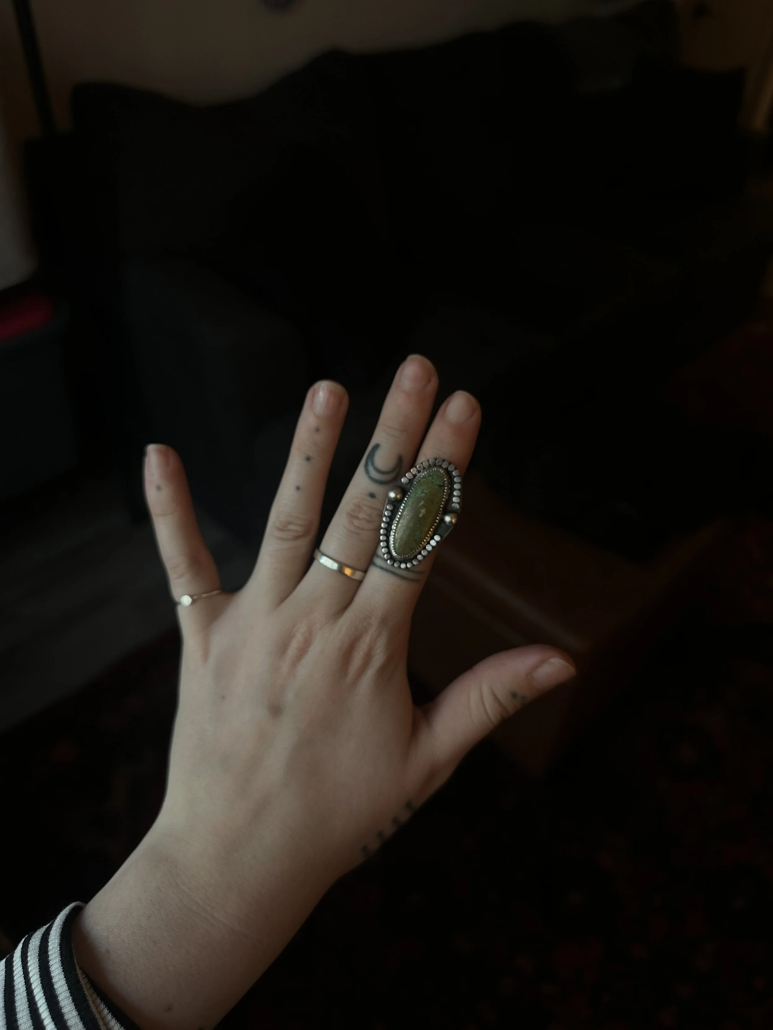 Close-up of a person's hand with jewelry and tattoos, against a dark background, with another person's arm wearing a striped sleeve visible in the foreground.