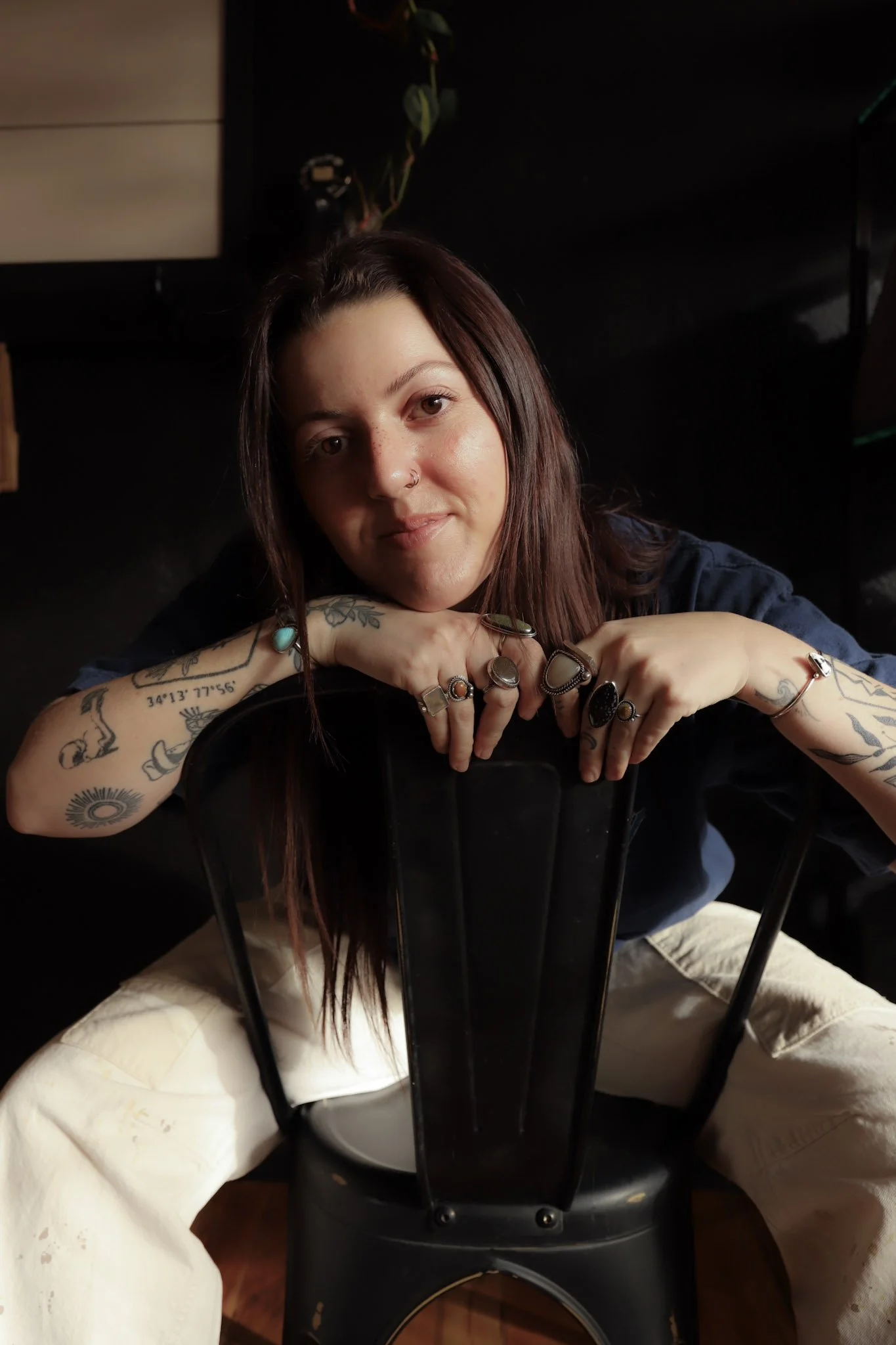 A woman with dark brown hair, tattoos on her arms, and multiple rings on her fingers, resting her chin on her hands while sitting behind a black chair with vertical bars in the backrest. She wears a dark top and light-colored pants.