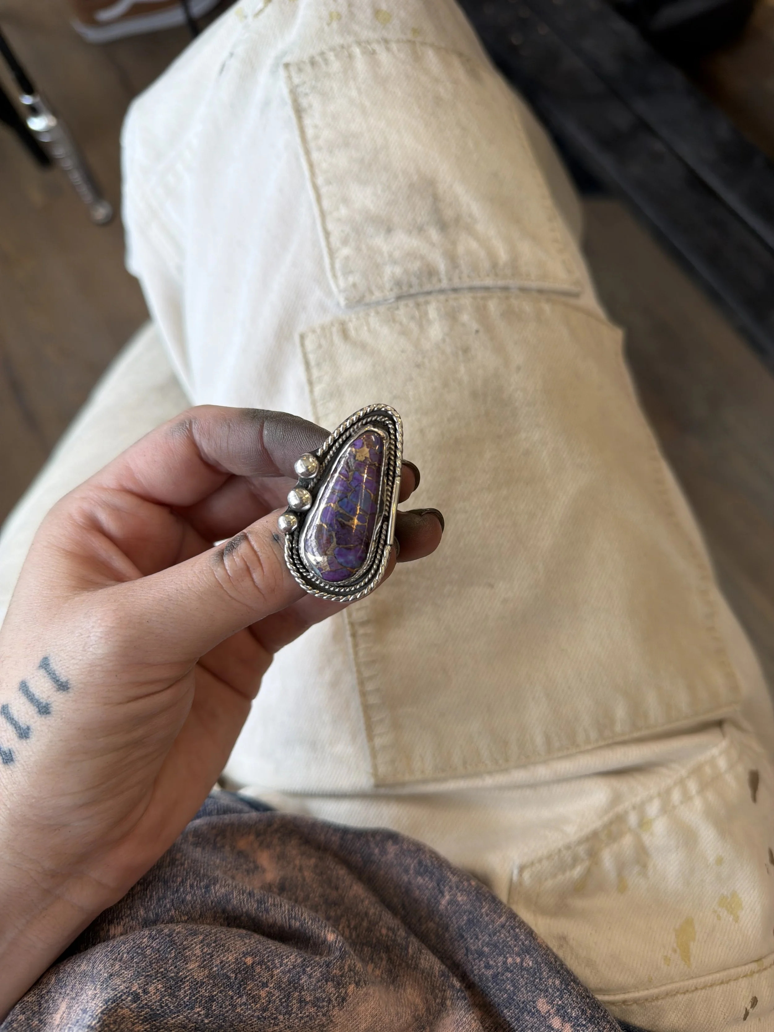 A person holding a large, ornate ring with purple and gold stones, sitting on a beige cloth or garment.