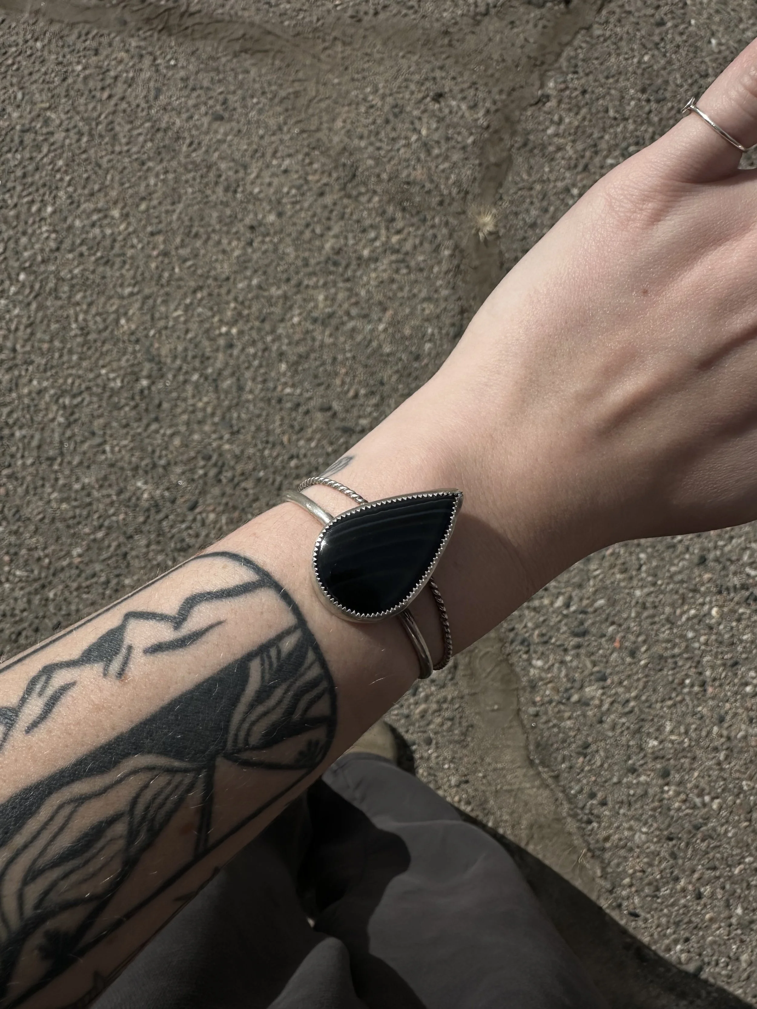 A person's arm and hand with tattoos, rings, and a black and silver bracelet, extending downward against a gravel ground surface.