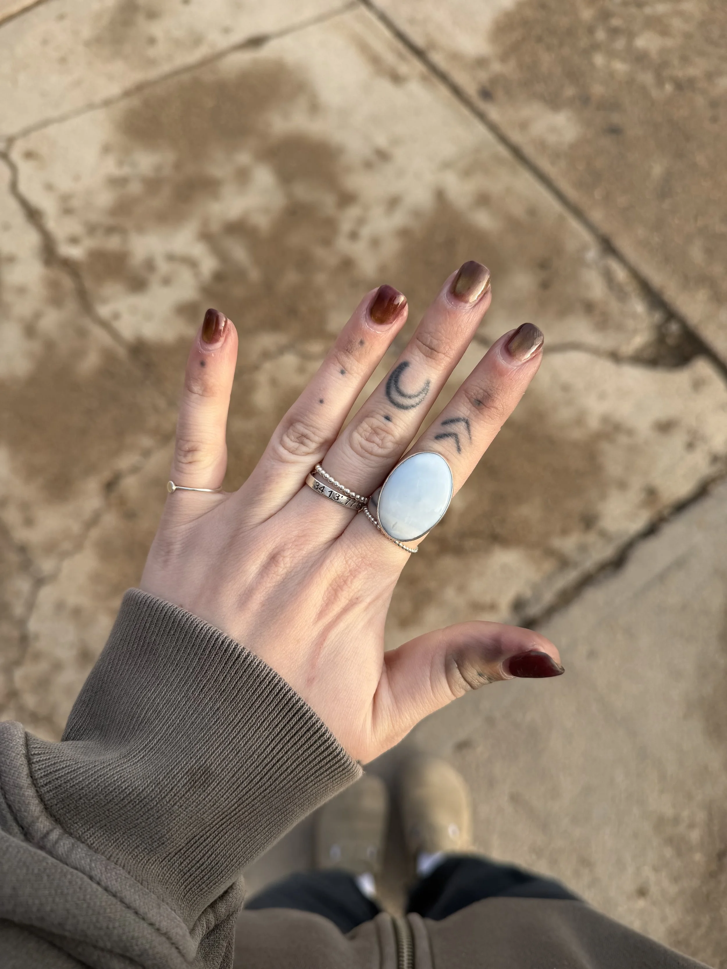 A person wearing a brownish gray sweater with their hand extended, showing ringed fingers with rose gold and silver rings and a tattooed middle finger, against a tiled floor background.