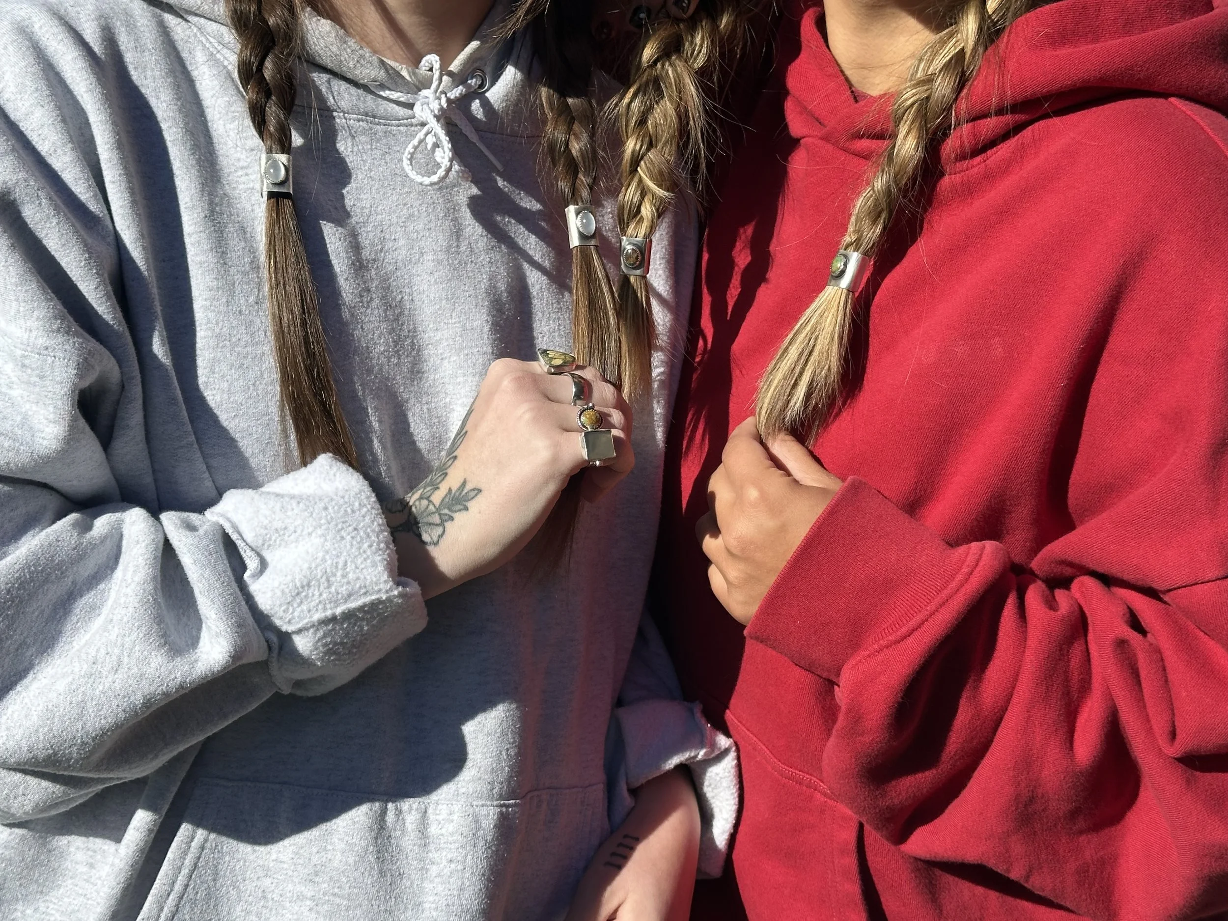 Two women standing close together, both wearing hoodies, one gray and one red, with braided hair and rings on their fingers.