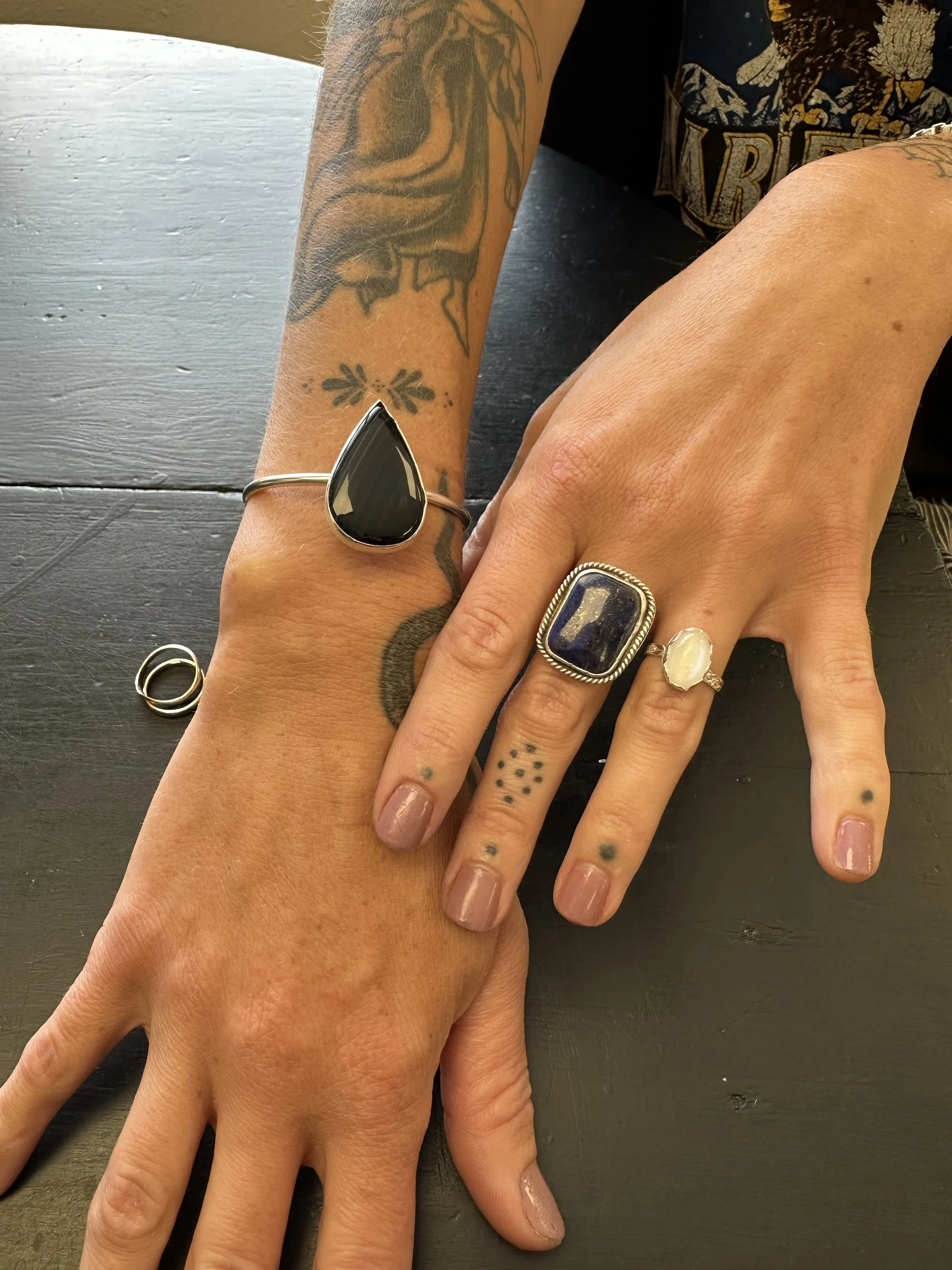Close-up of two hands with rings resting on a dark wooden surface. The left hand has a black stone ring, and the right hand has a large purple gemstone ring, along with a smaller pearl ring. The hands have small tattoos and are adorned with multiple 