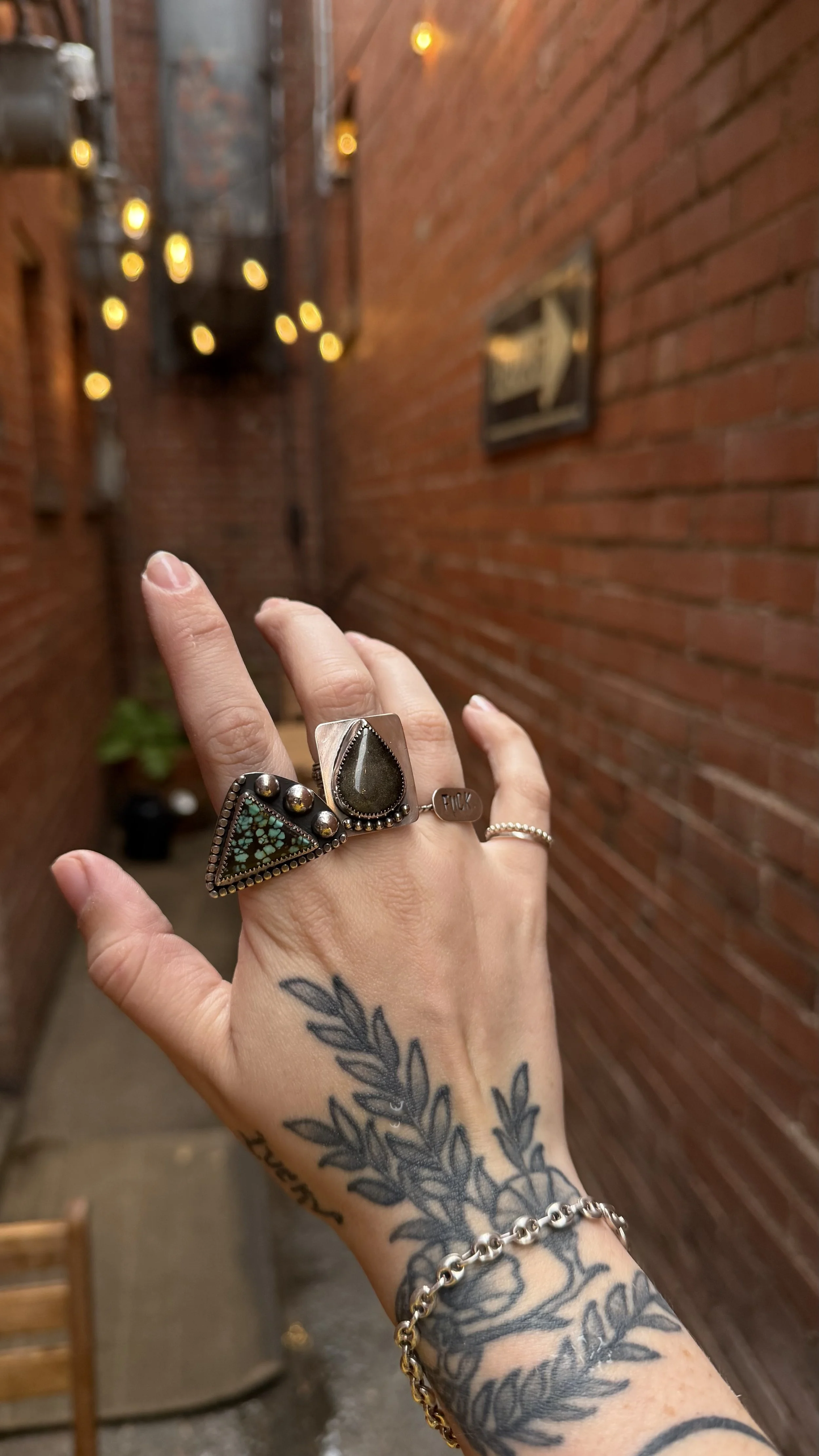 A tattooed hand wearing multiple rings and a bracelet, held in front of a red brick wall with blurred warm light bulbs and framed artwork in the background.
