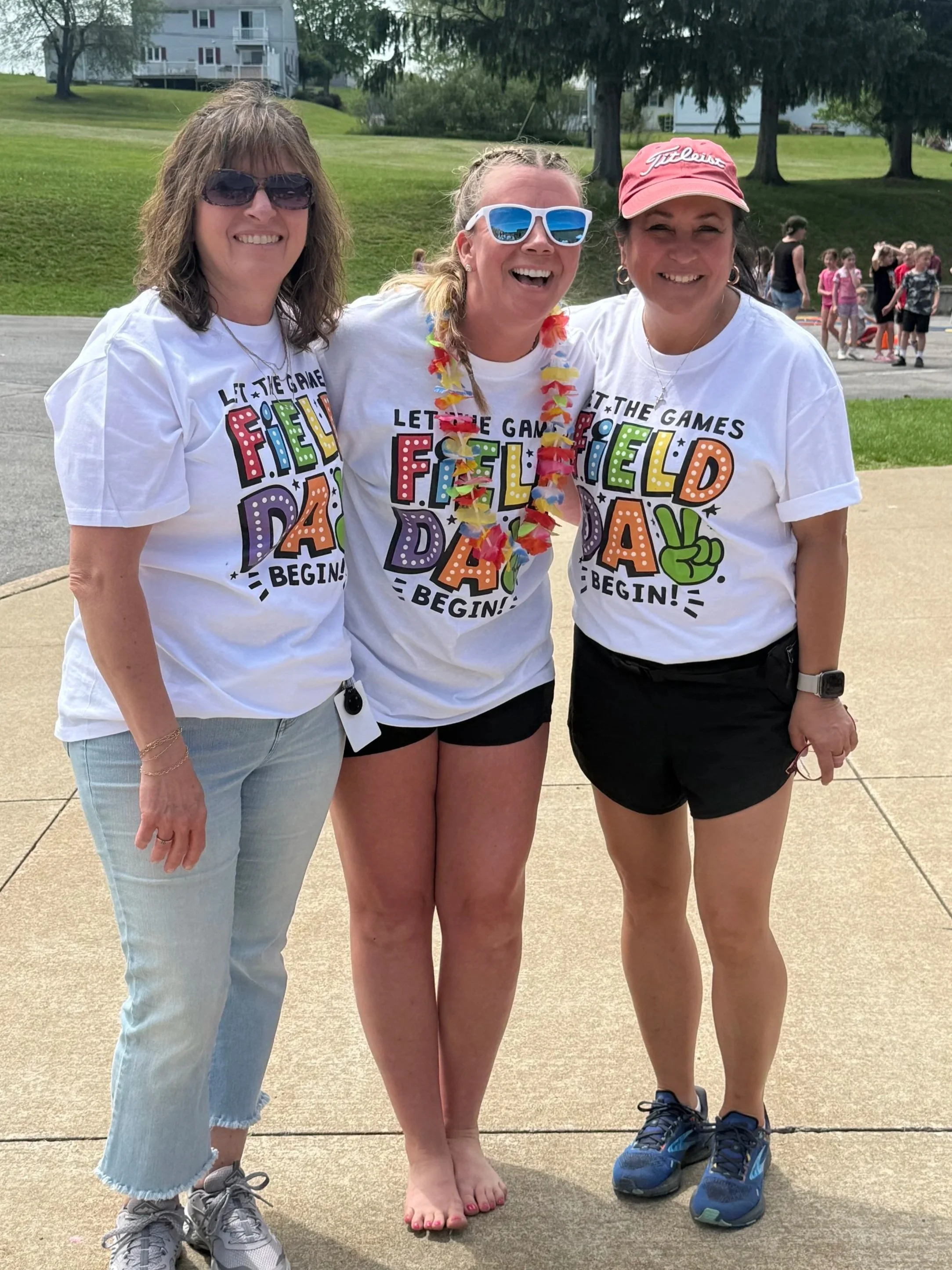 Three women standing close together outdoors, smiling, wearing casual clothes and sunglasses. The woman in the middle is barefoot with her heels off and wearing a colorful lei, while the other two women are wearing sneakers. In the background, a group of children and some adults are gathered, with trees and a grassy hill visible.