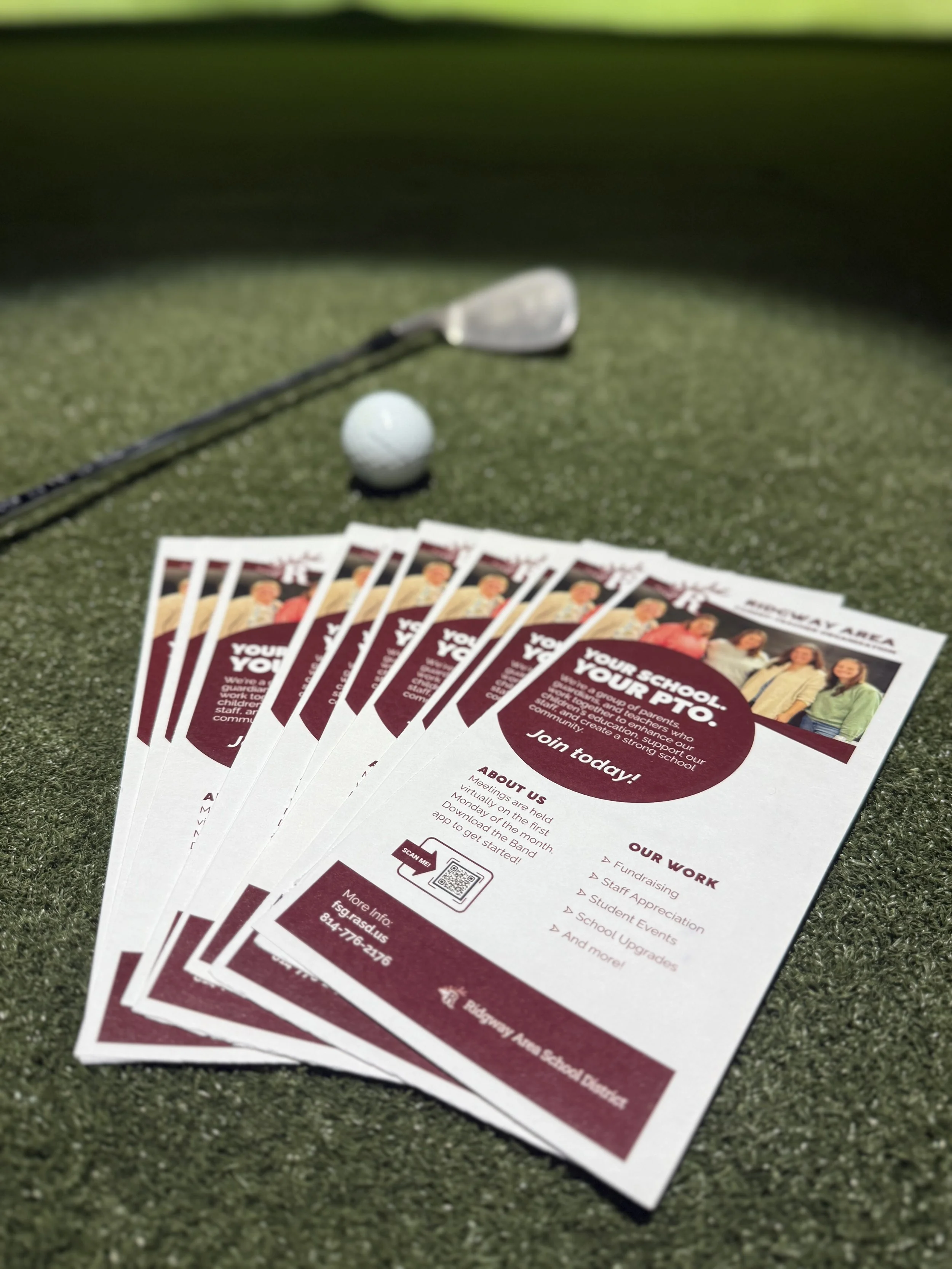 Flyers for a school event, laid out on green turf, with a golf club and golf ball in the background.
