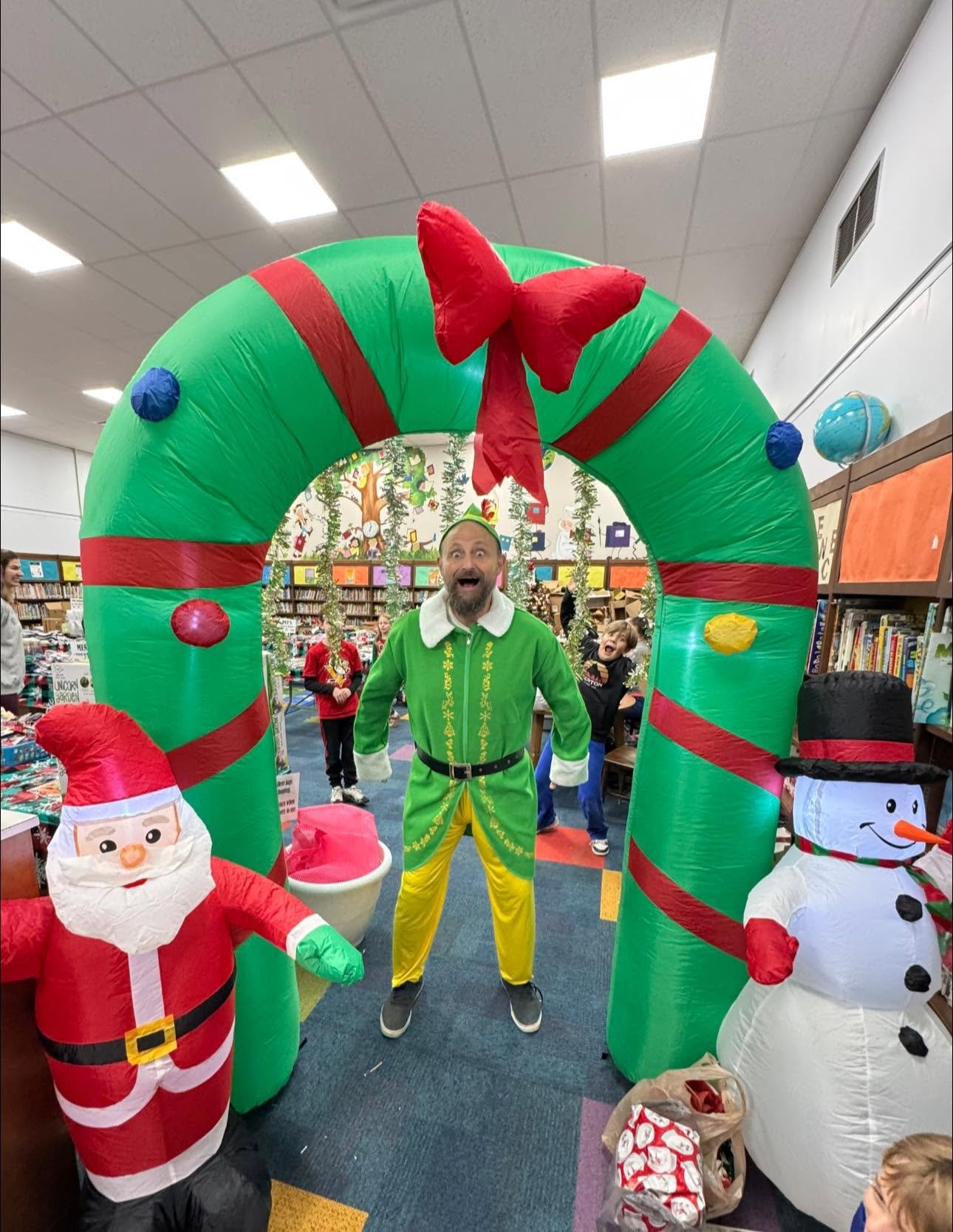 A man dressed as an elf standing under a large inflatable Christmas candy cane arch in a festive event at a library. Children are in the background, with inflatable Santa and snowman decorations nearby.