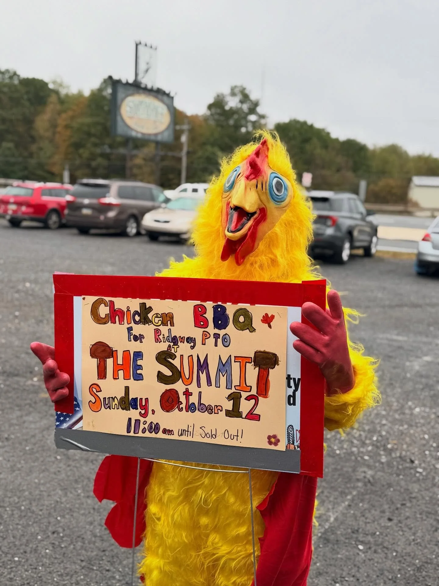 Person dressed in a chicken costume holding a colorful sign advertising a chicken BBQ event on Sunday, October 12 from 11:00 am until sold out. The background shows a parking lot with cars and trees.