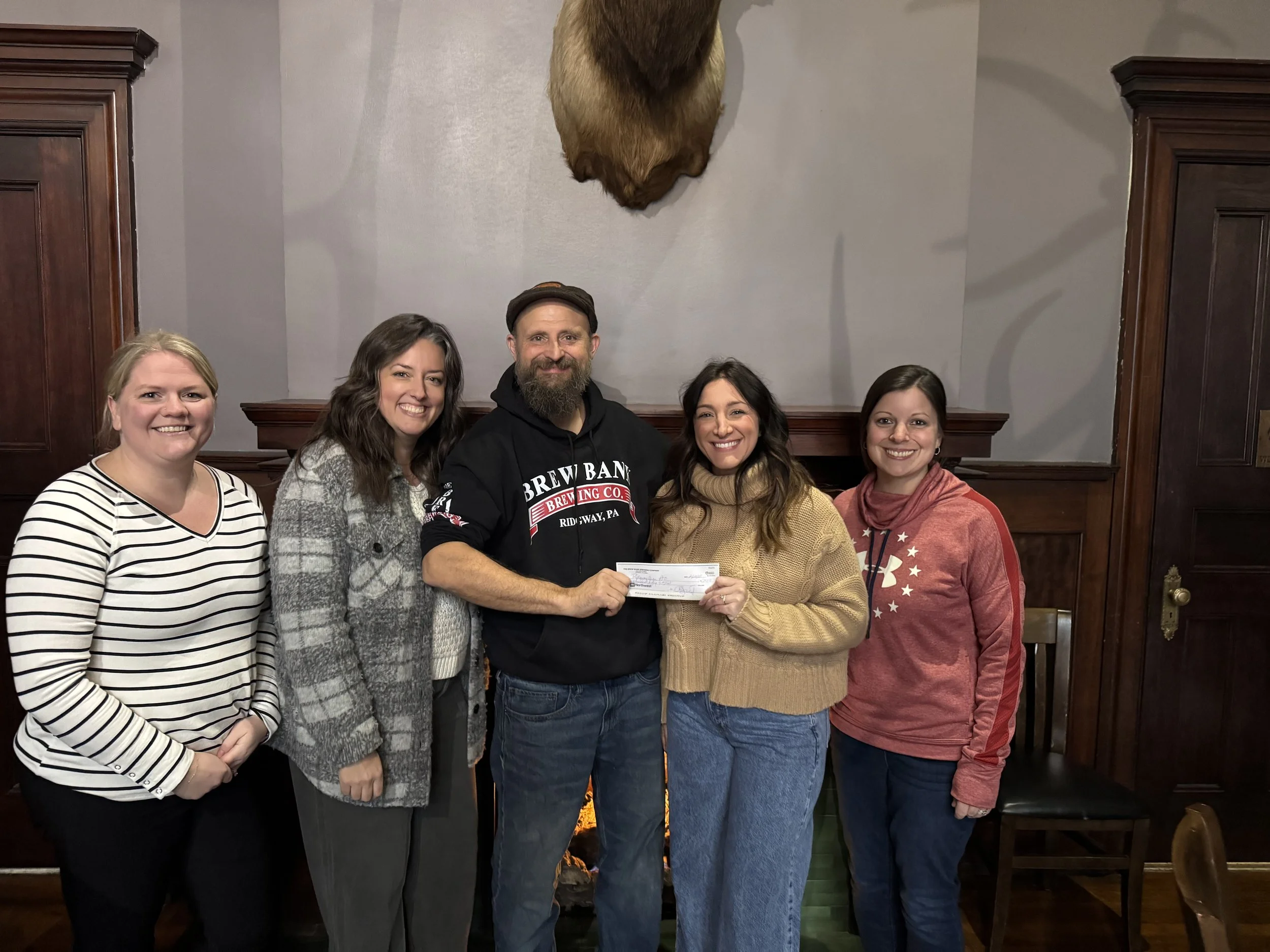 A group of five smiling people standing in front of a grey wall with a mounted bear's head in a wood-paneled room. One person is holding a check.