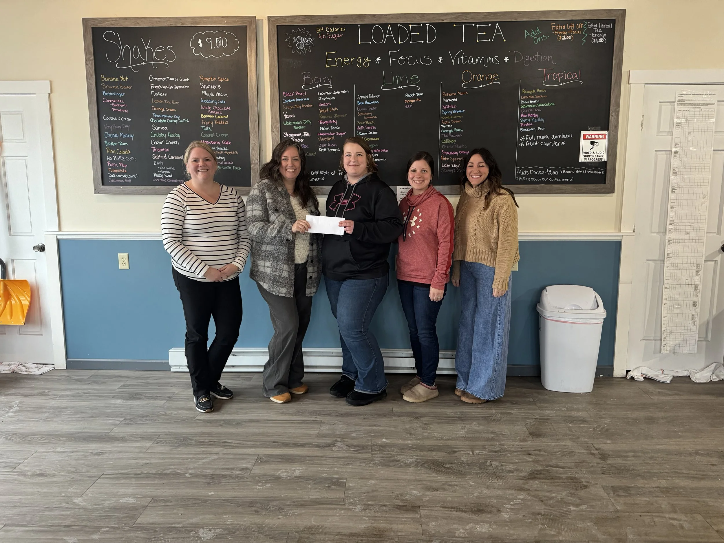 Five women standing together in front of a large blackboard menu inside a cafe, smiling at the camera. The blackboard has colorful handwritten menu items including shakes and loaded teas.