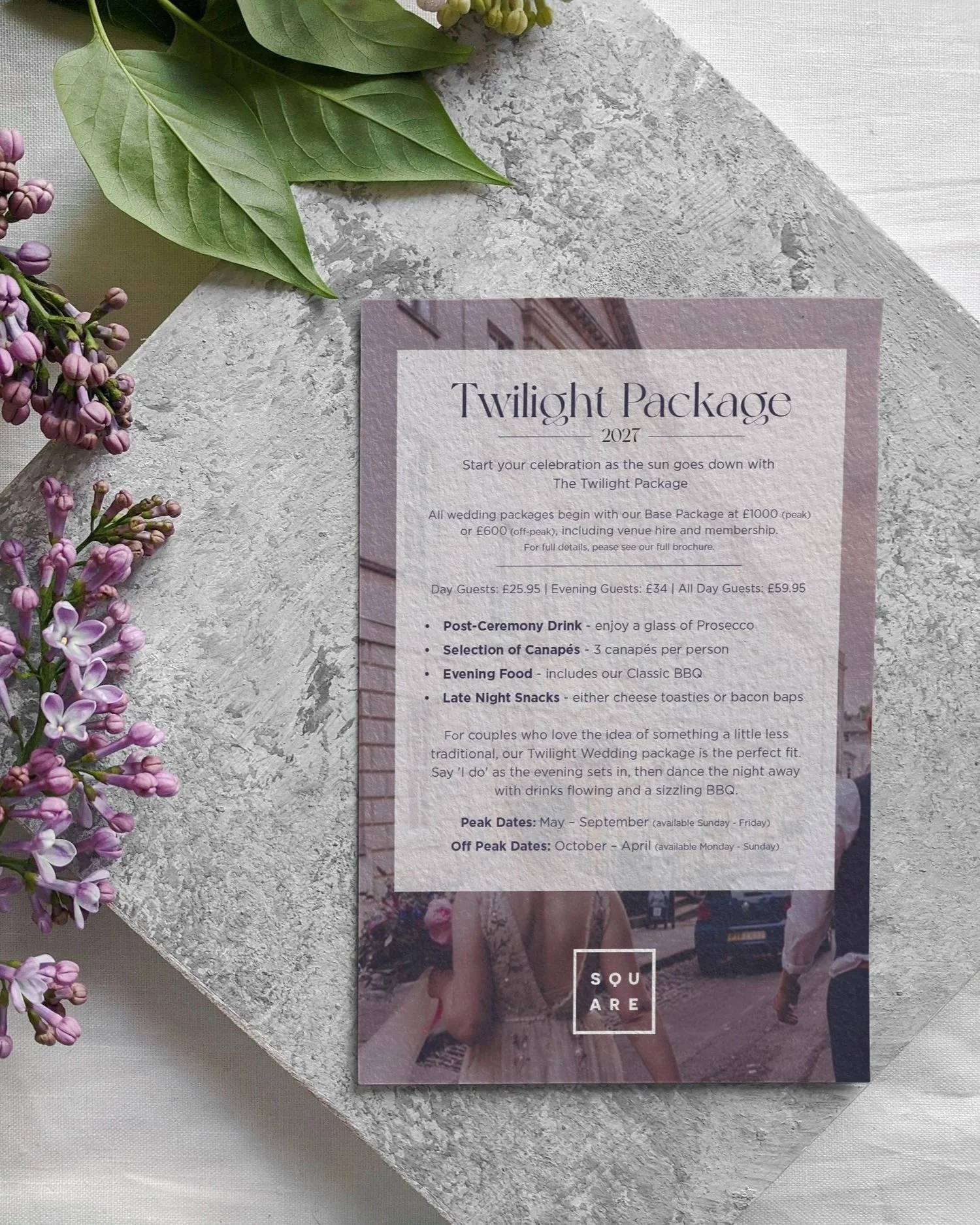 A wedding brochure titled "Twilight Package" is placed on a textured gray surface, surrounded by purple lilac flowers and green leaves. The brochure provides details about wedding packages and celebration options.