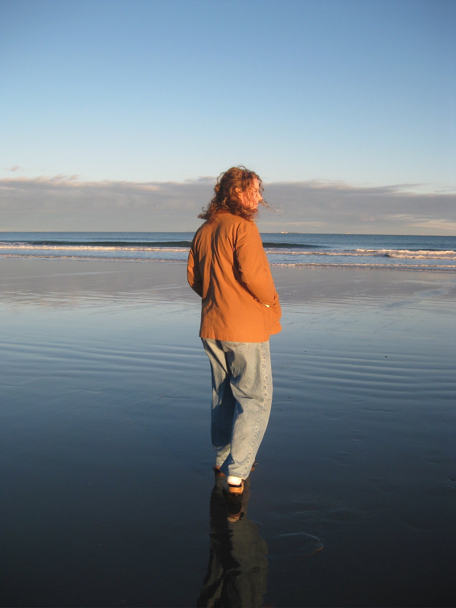 A person with curly hair wearing a brown jacket and light jeans stands on wet sand at the beach, facing sideways with their hands in their pockets, while the ocean and sky are visible in the background during the daytime.