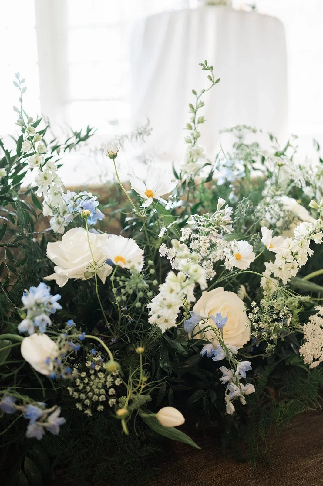 A floral arrangement with white roses, daisies, and various white and blue flowers surrounded by greenery on a dark surface, with a bright window in the background.