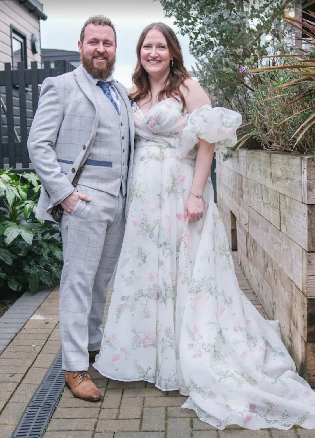 A man and woman dressed in formal attire, standing outdoors on a brick pathway next to a wooden planter with greenery. The man has a beard, wearing a light gray checkered suit with a blue tie and brown shoes. The woman has long hair, wearing a white floral dress with puffed sleeves.