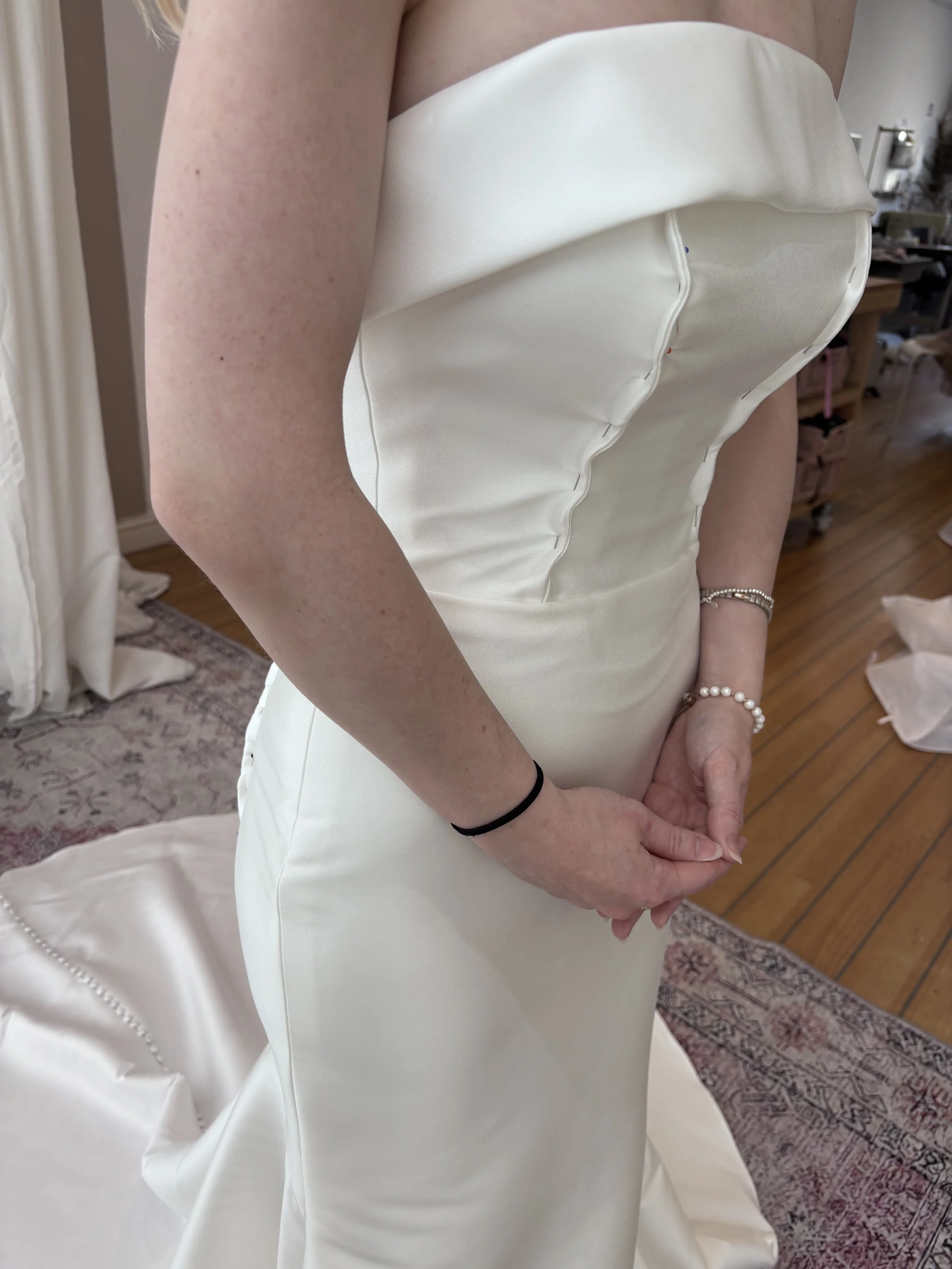 Close-up of a person in a wedding dress, showing their arm, part of the dress, and jewelry on their wrist.