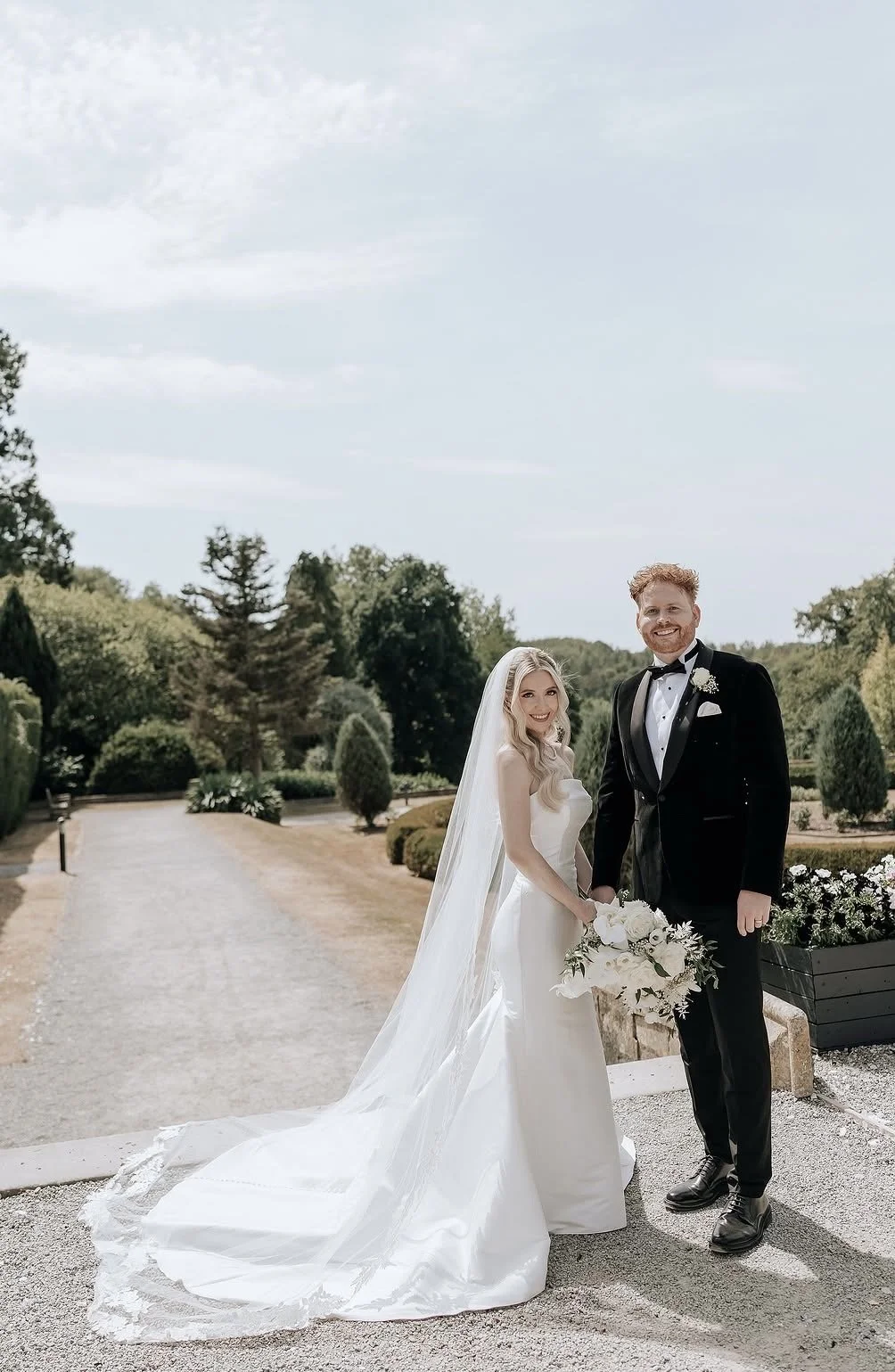 A bride and groom holding hands outdoors on a wedding day. The bride is wearing a white wedding gown with a long train and veil, and is holding a bouquet of white flowers. The groom is dressed in a black tuxedo with a bow tie, and is smiling. The background features a landscaped garden with trees and a clear sky.